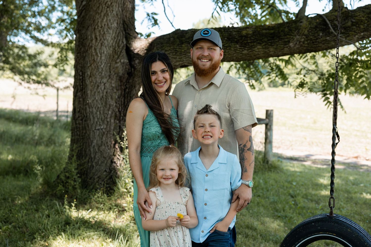 Happy family of four posing together under a large tree near a tire swing in a sunny field.