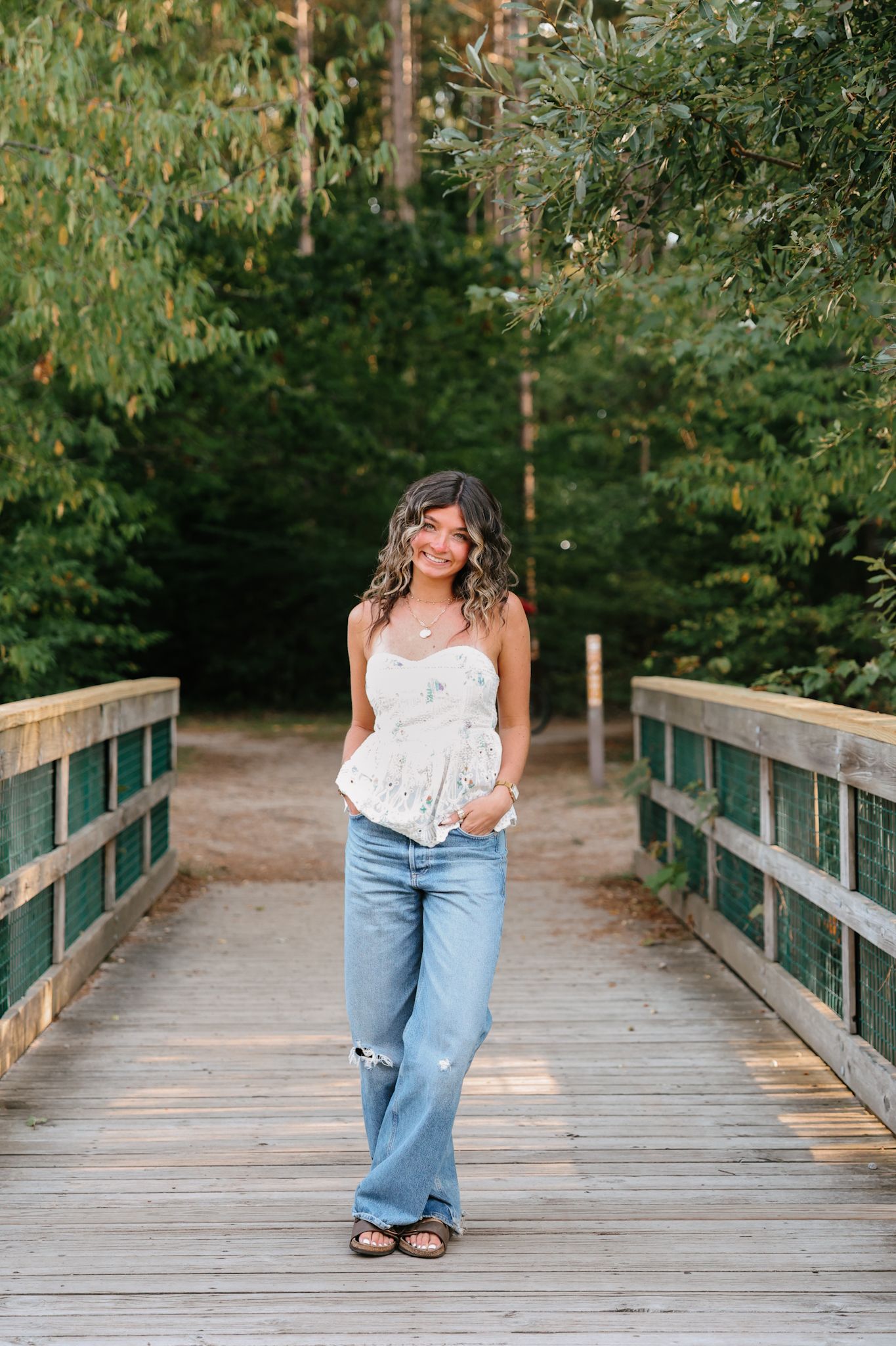 Young woman in lace top and jeans posing on wooden bridge in woods.