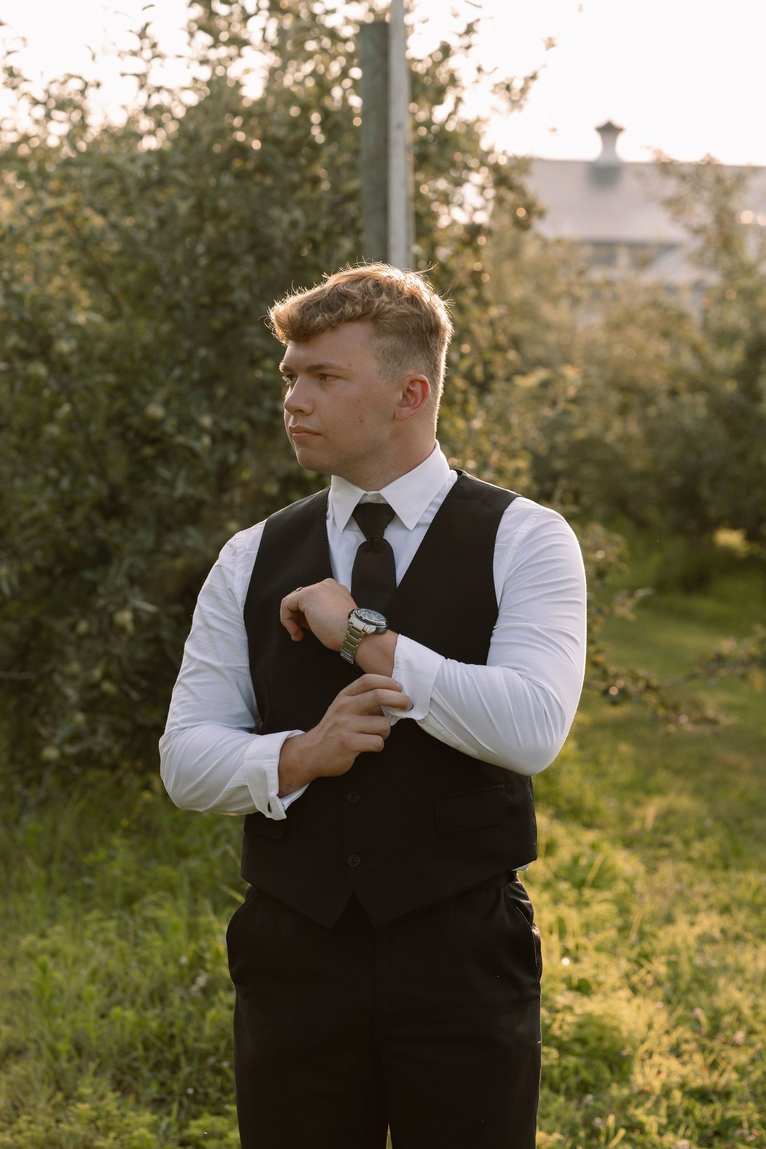 Dapper groom in black vest and tie adjusting cufflinks outdoors during golden hour wedding photos.