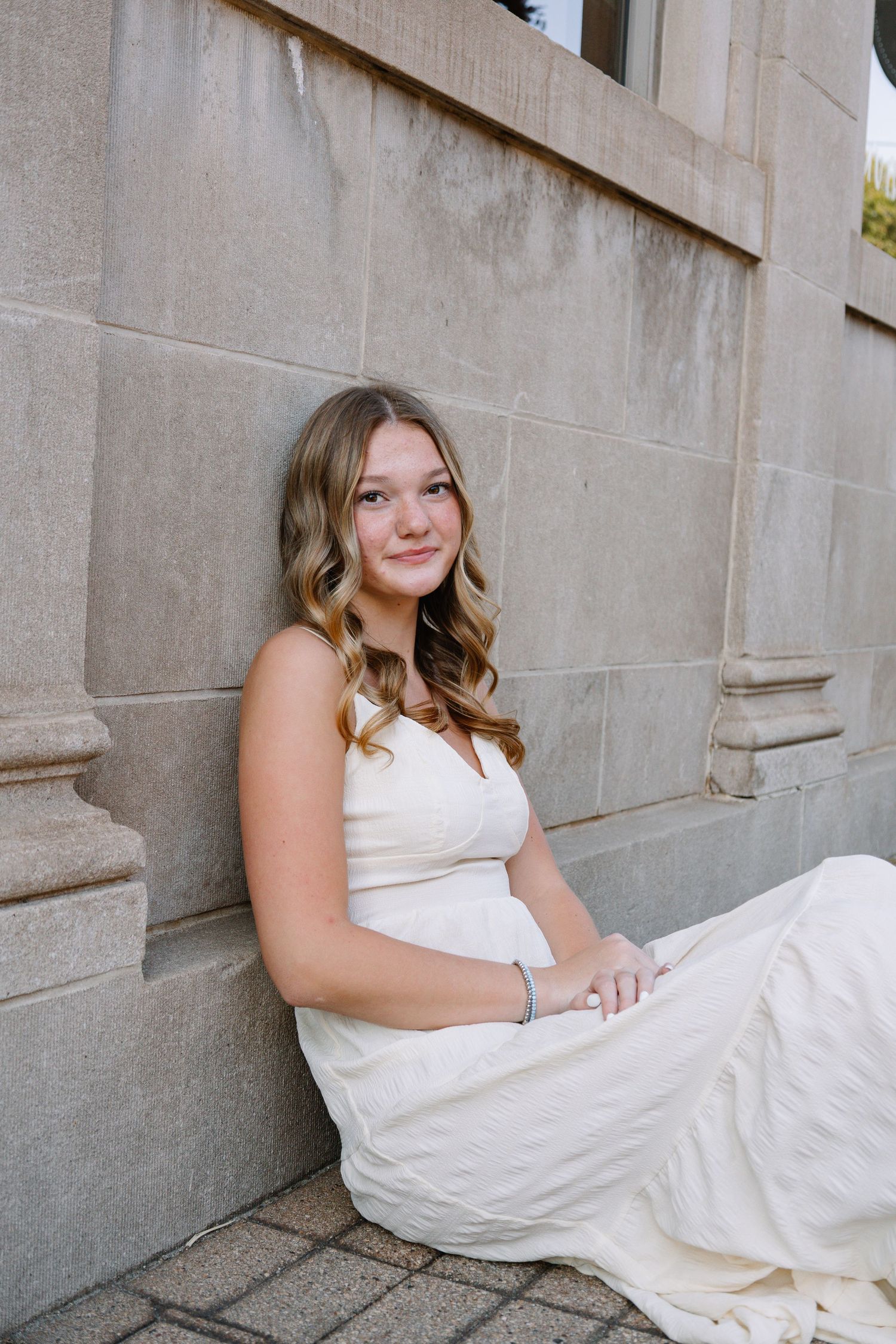 Young woman in white dress sitting against stone wall, smiling softly in elegant outdoor portrait.