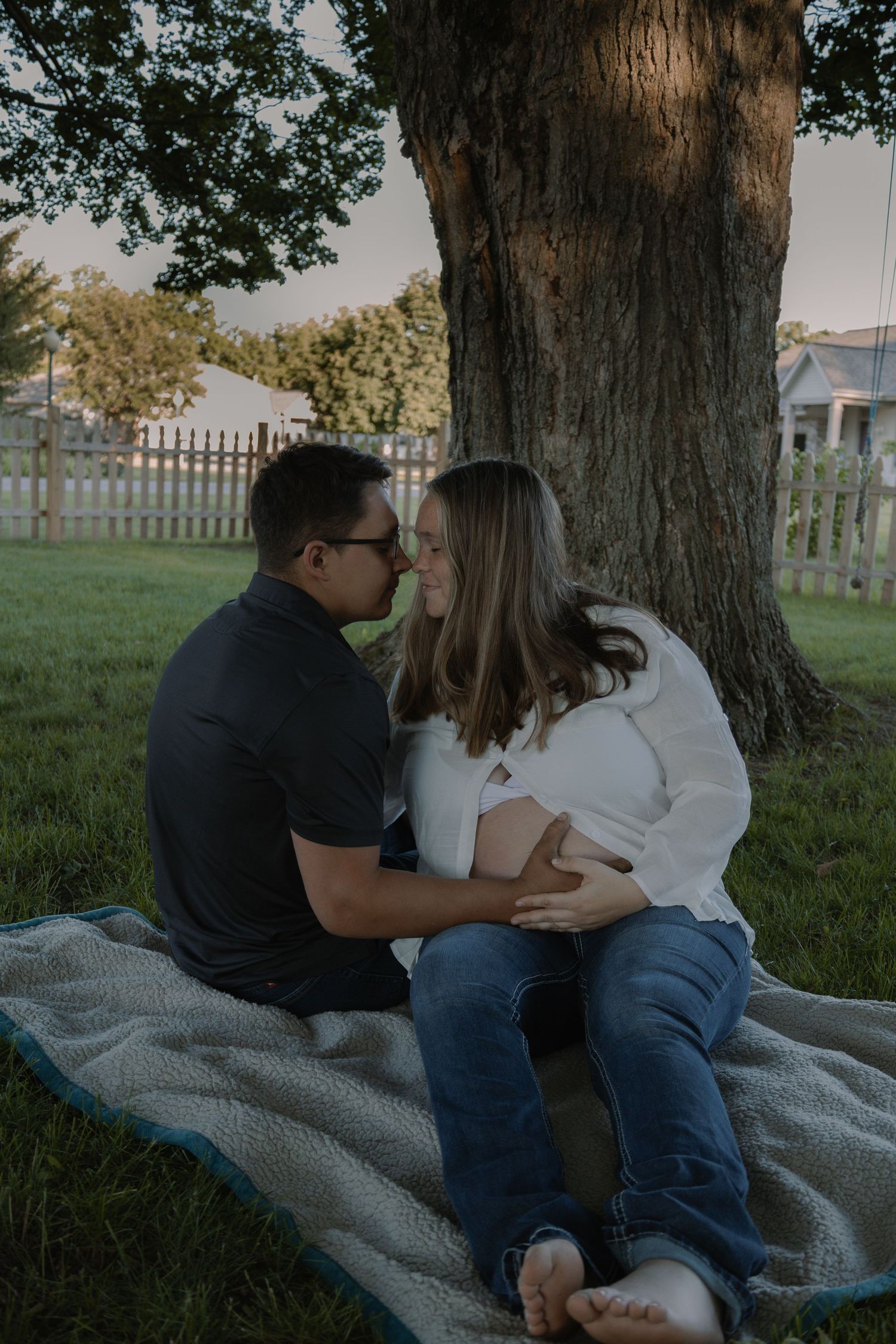Pregnant couple sitting on blanket under large tree, sharing a romantic nose-to-nose moment outdoors.