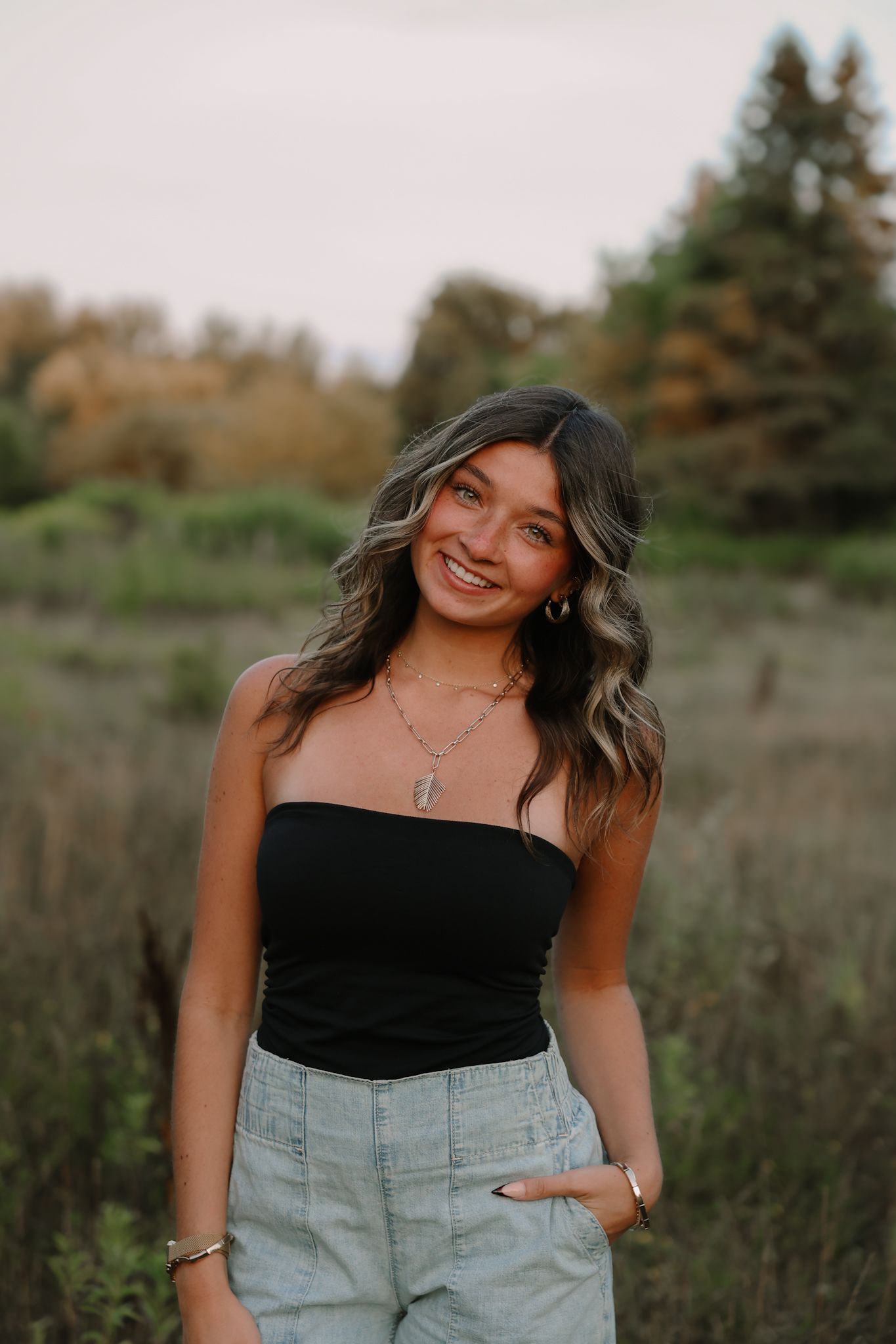 Smiling brunette woman in black strapless top and light jeans posing in autumn field.