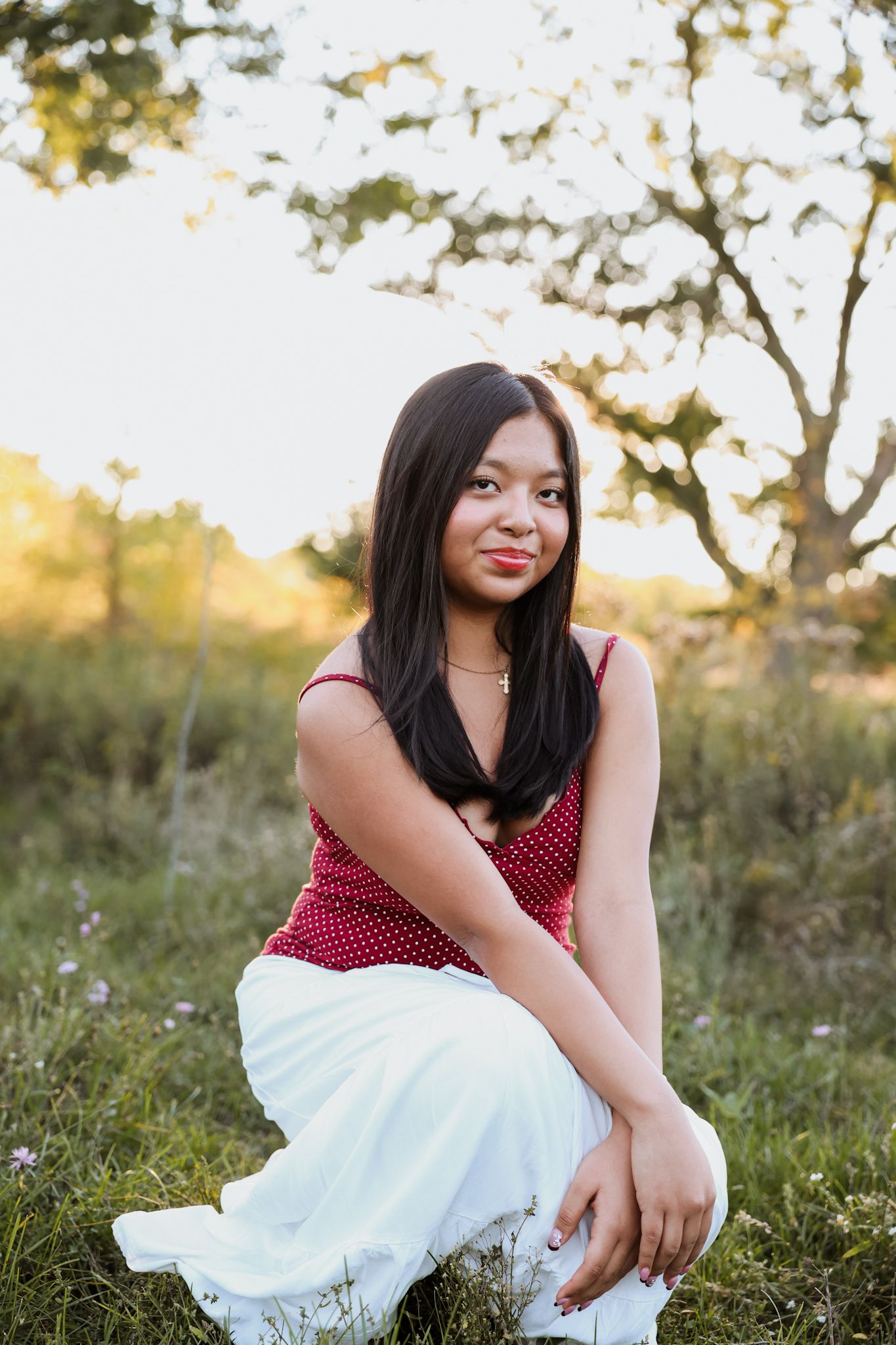 Young Asian woman in red polka dot top and white skirt sits in a sunlit grassy meadow.