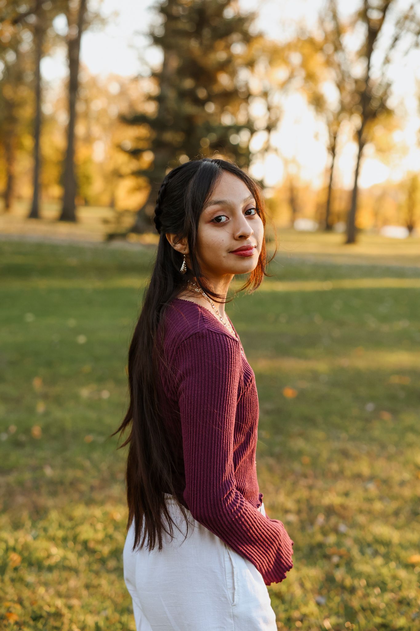 Young woman with long dark hair in burgundy sweater posing in golden autumn park at sunset.