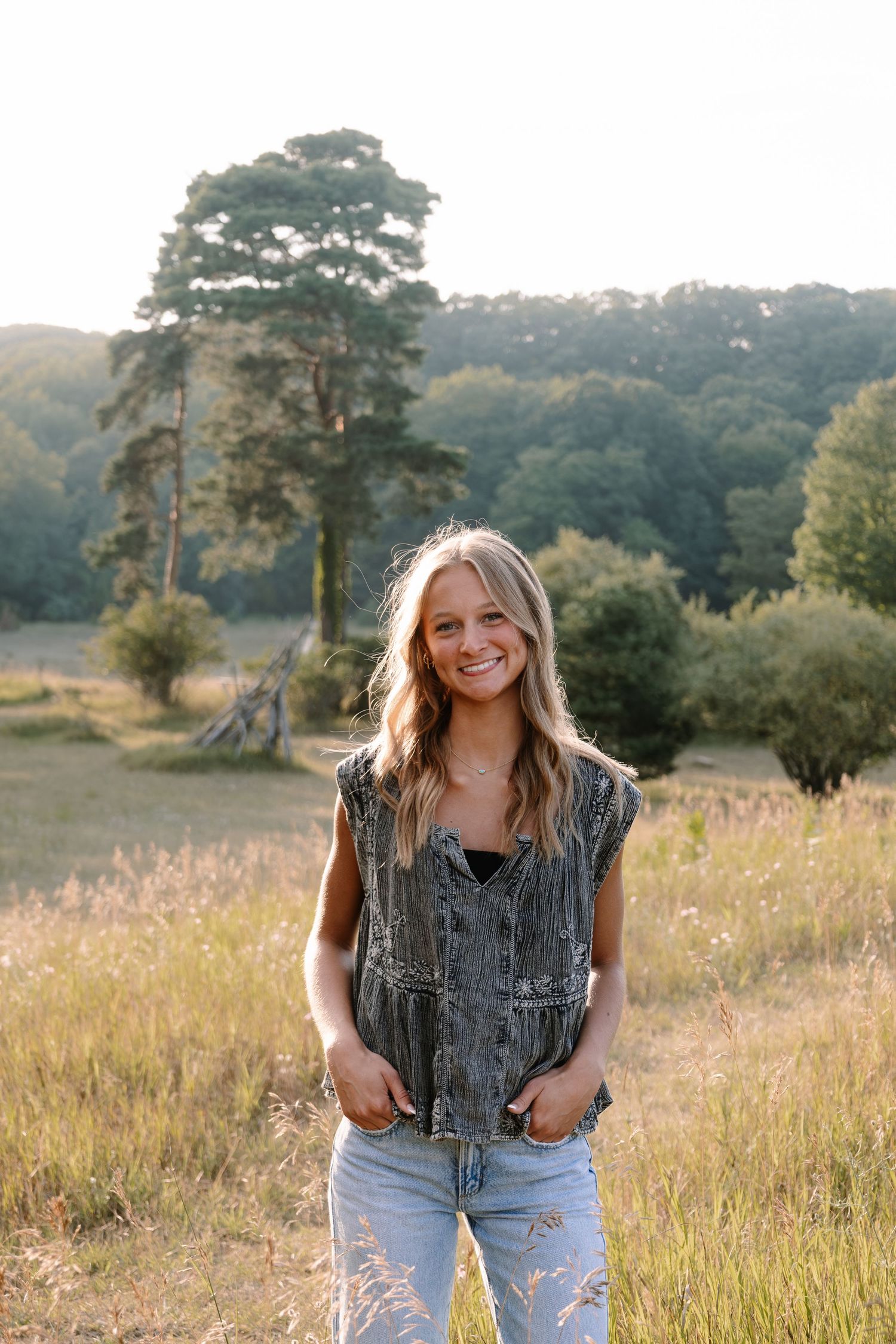 Smiling blonde woman standing in a sunlit meadow wearing a gray denim vest and light jeans with trees in the background.