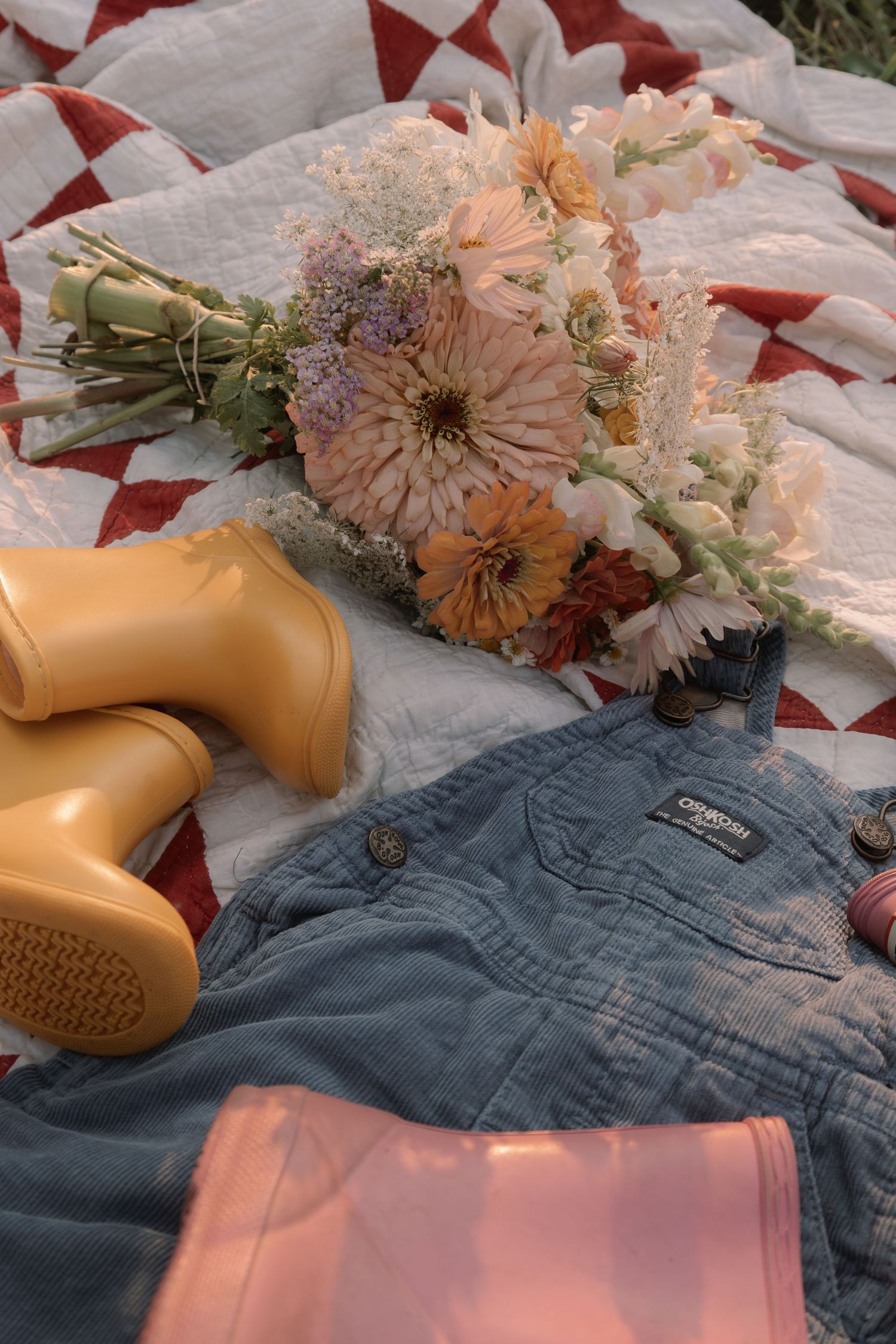 Flat lay of yellow rubber boots, wildflower bouquet, denim overalls on red and white quilt outdoors.