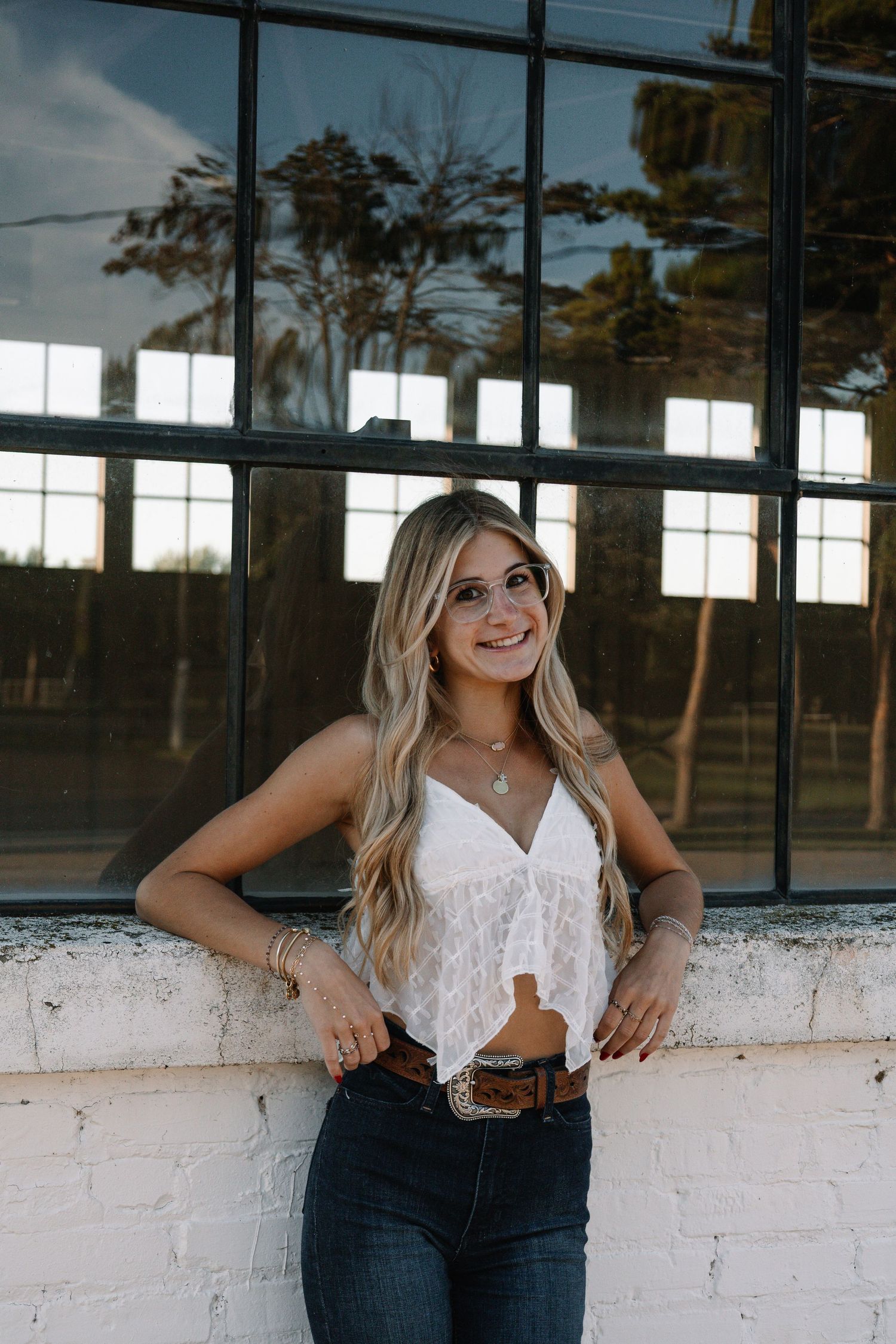 Young blonde woman in white lace top and jeans smiling outdoors against a brick wall and large windows.