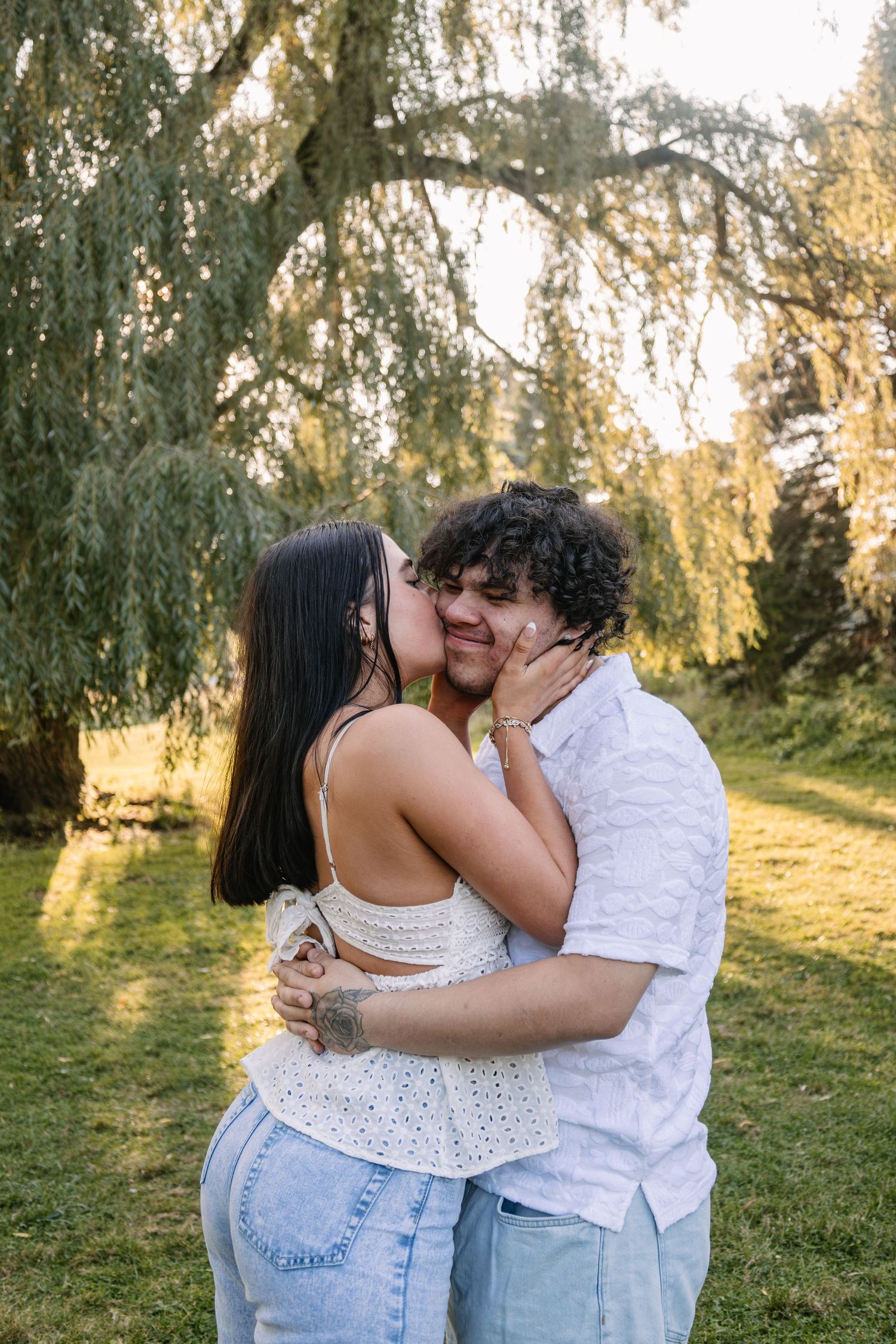 A couple shares a romantic kiss under a willow tree in a sunlit park, woman in white top and jeans.