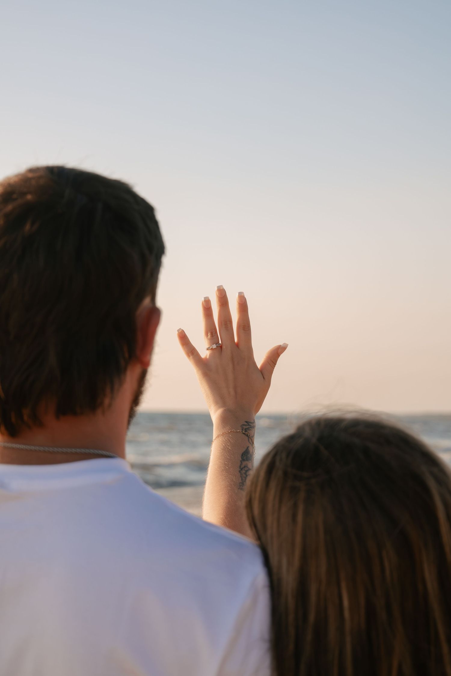 Couple at beach sunset, woman showing engagement ring with floral wrist tattoo, ocean horizon in background.
