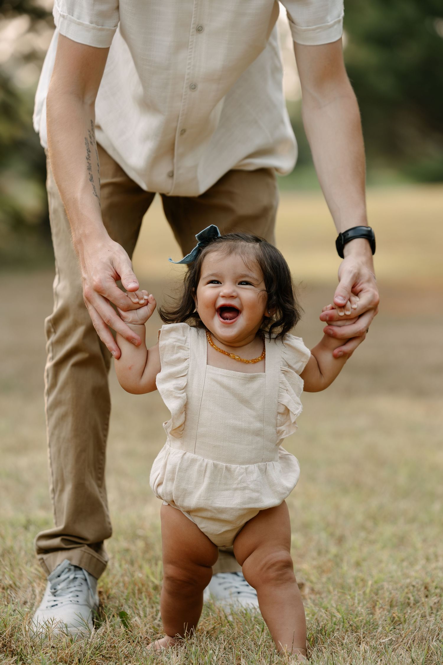 A joyful baby girl takes first steps outdoors, held by an adult's hands, wearing a cream ruffle romper with a bow headband.