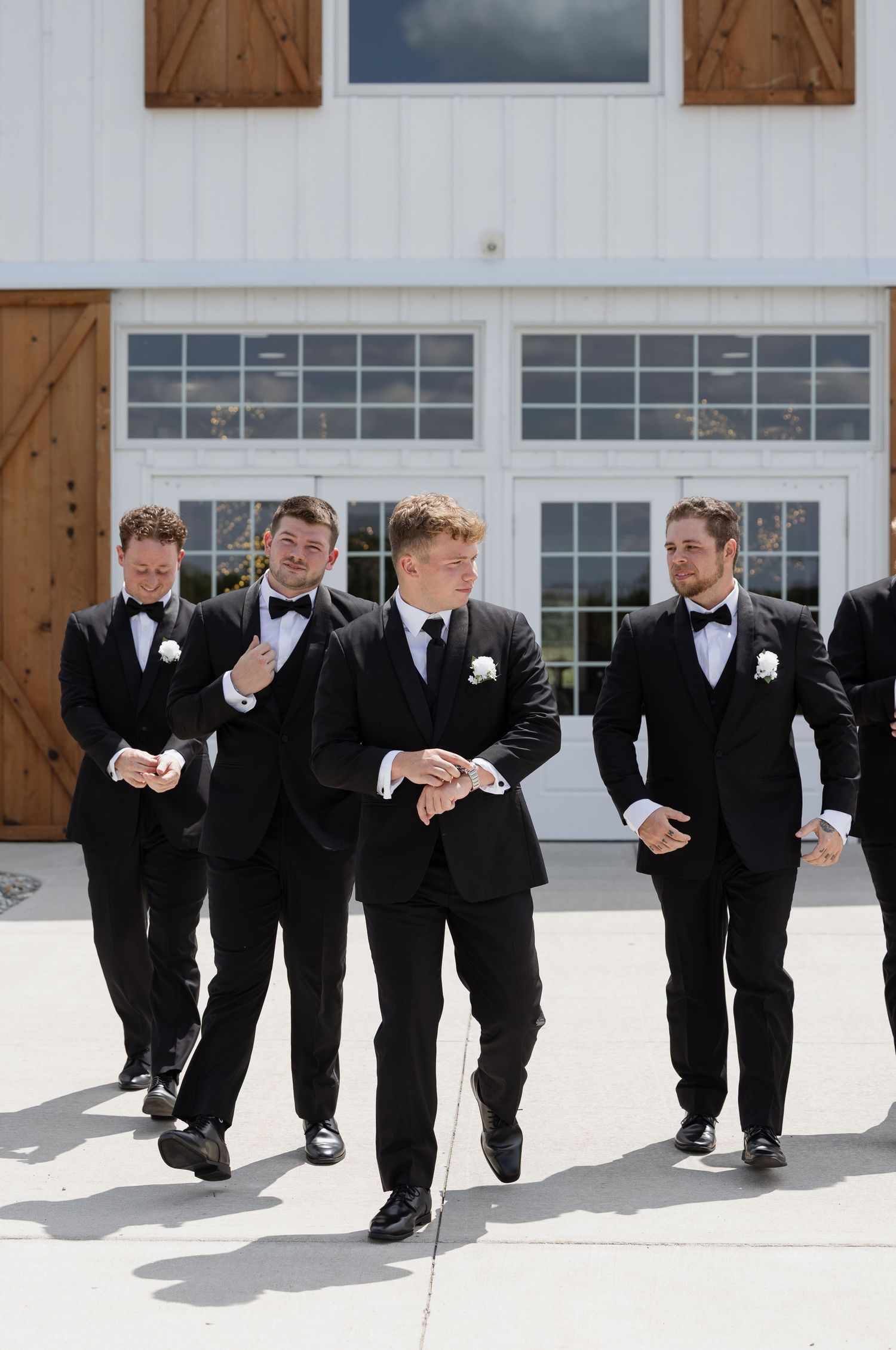 Four groomsmen in black tuxedos with bow ties and boutonnieres walk confidently outside a white barn wedding venue.