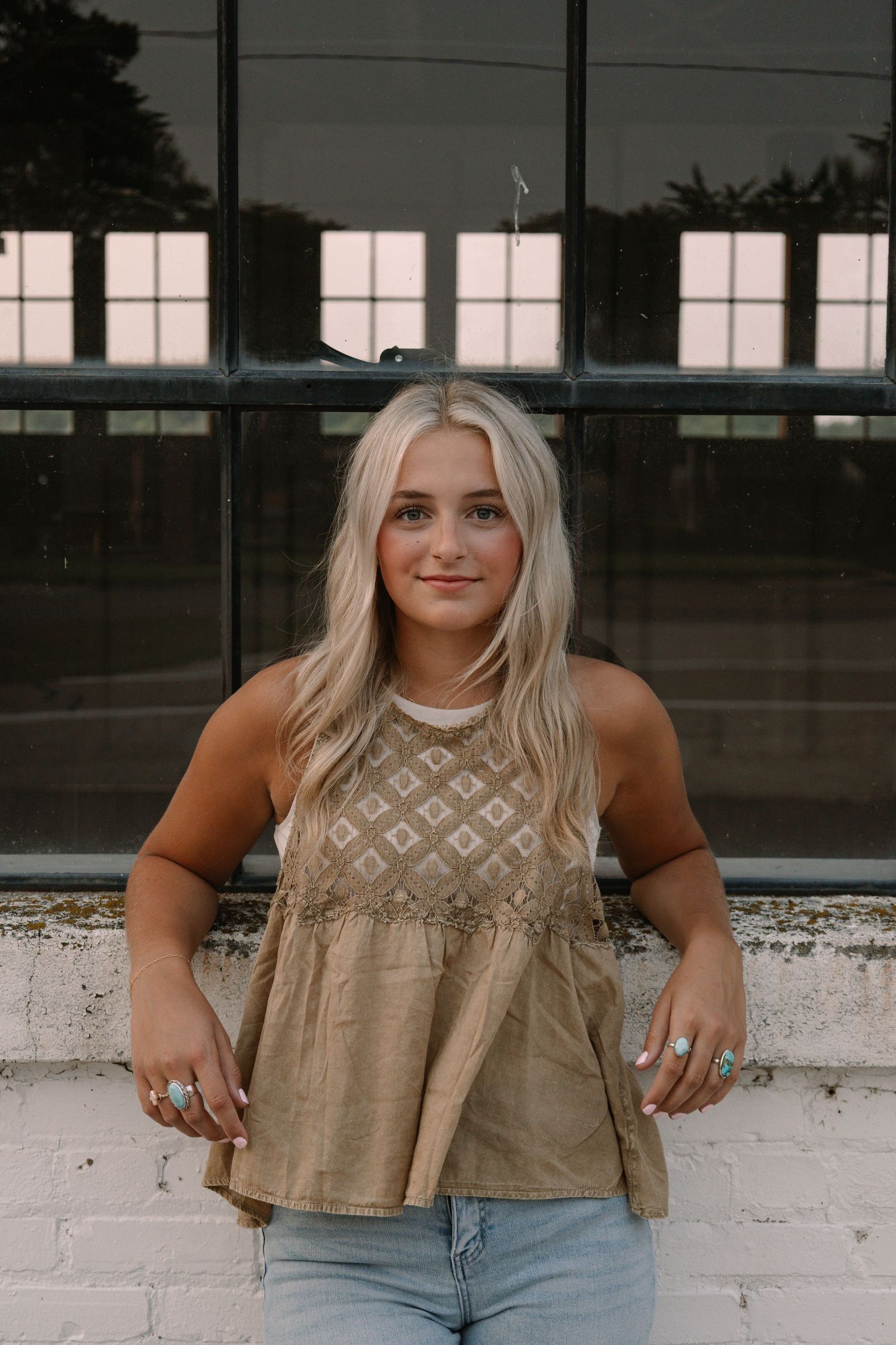 Young woman with blonde hair wearing a tan embroidered top and jeans, posing in front of large windows.
