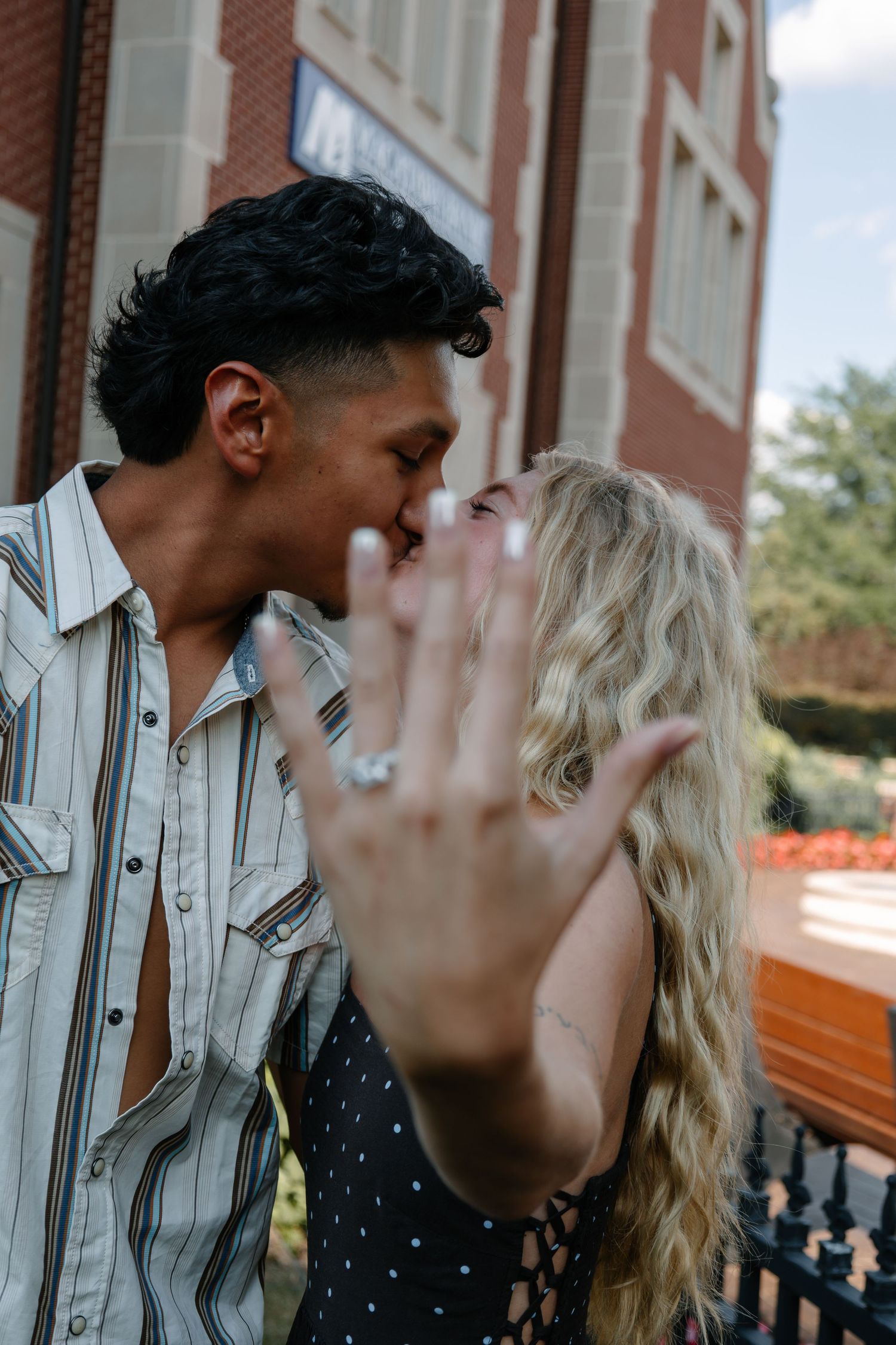 Couple kissing while showing off engagement ring, celebrating their proposal outdoors near a brick building.