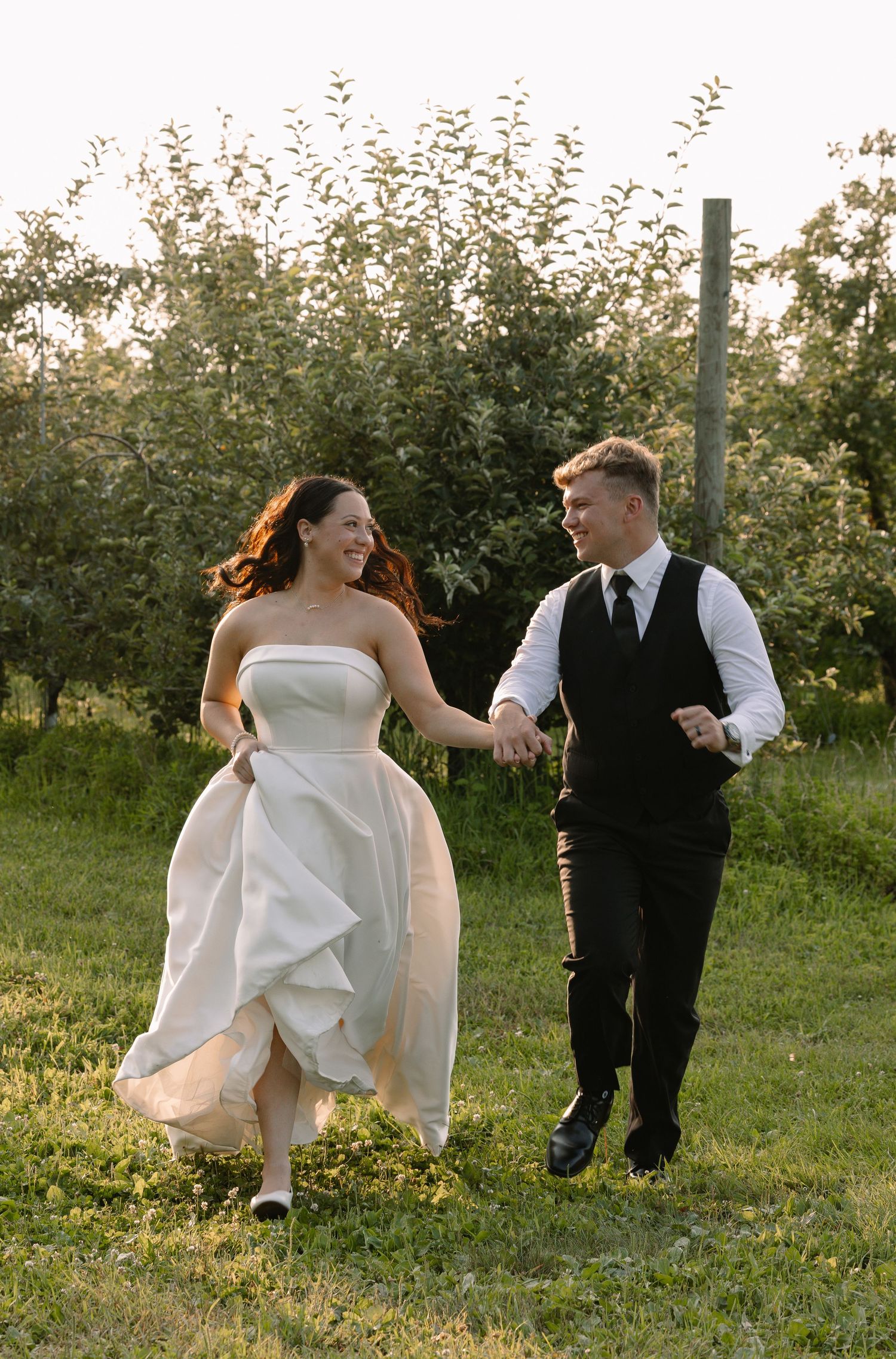 Joyful bride and groom run hand-in-hand through a sunlit orchard in wedding attire.