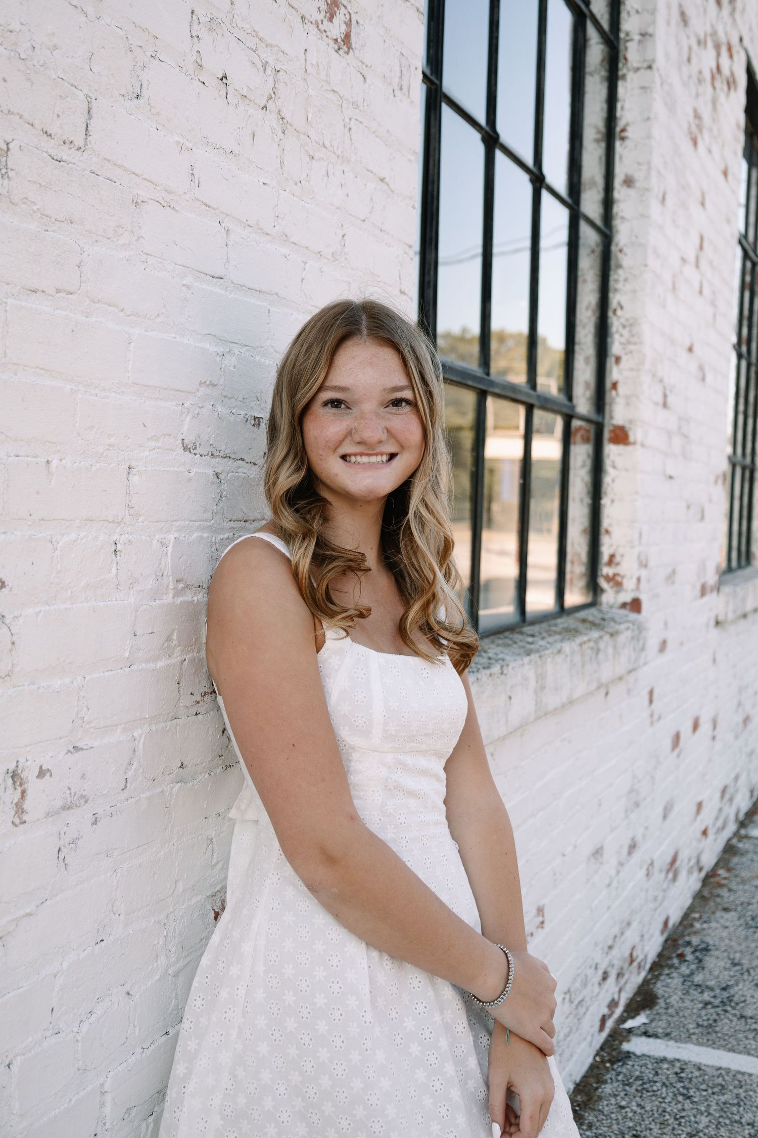 Young woman in white sundress smiling against white brick wall near industrial window, senior portrait photography.
