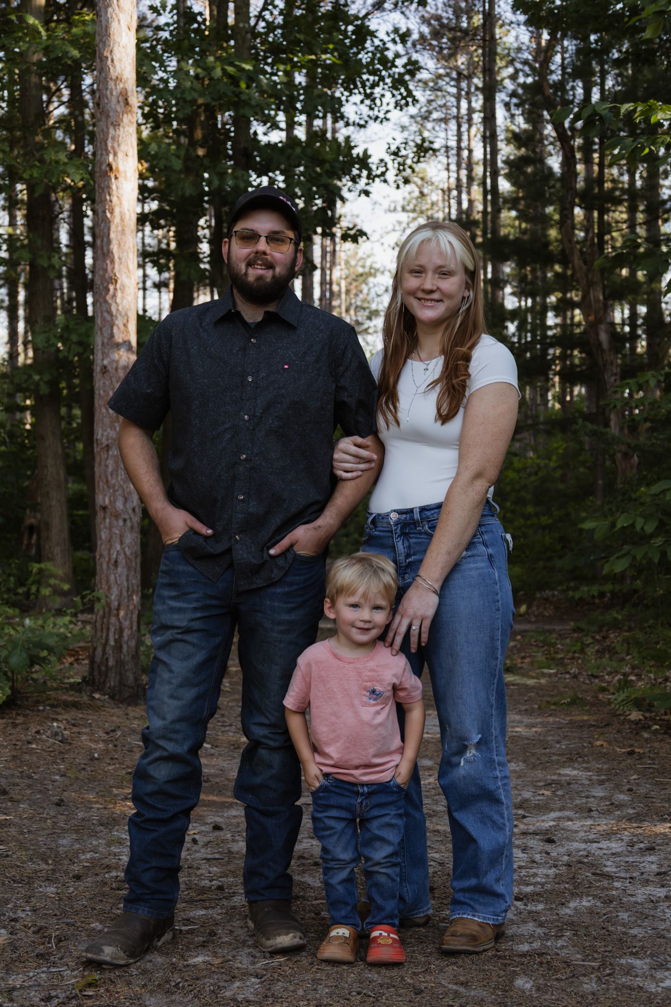 A family of three poses in a pine forest, with two adults and a young toddler smiling for a portrait photo.