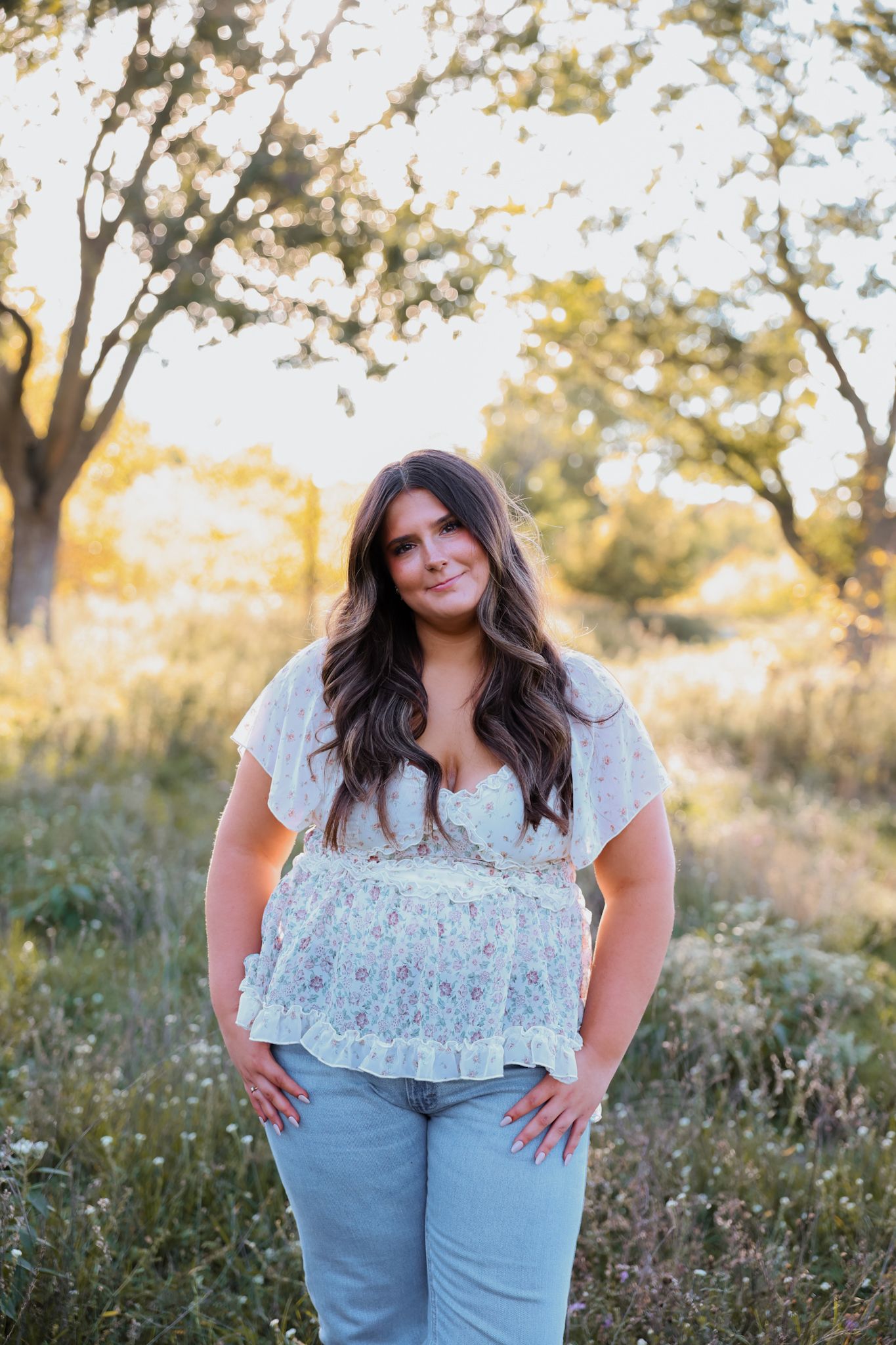 Young woman in white floral top and jeans standing on a sunlit path surrounded by greenery.
