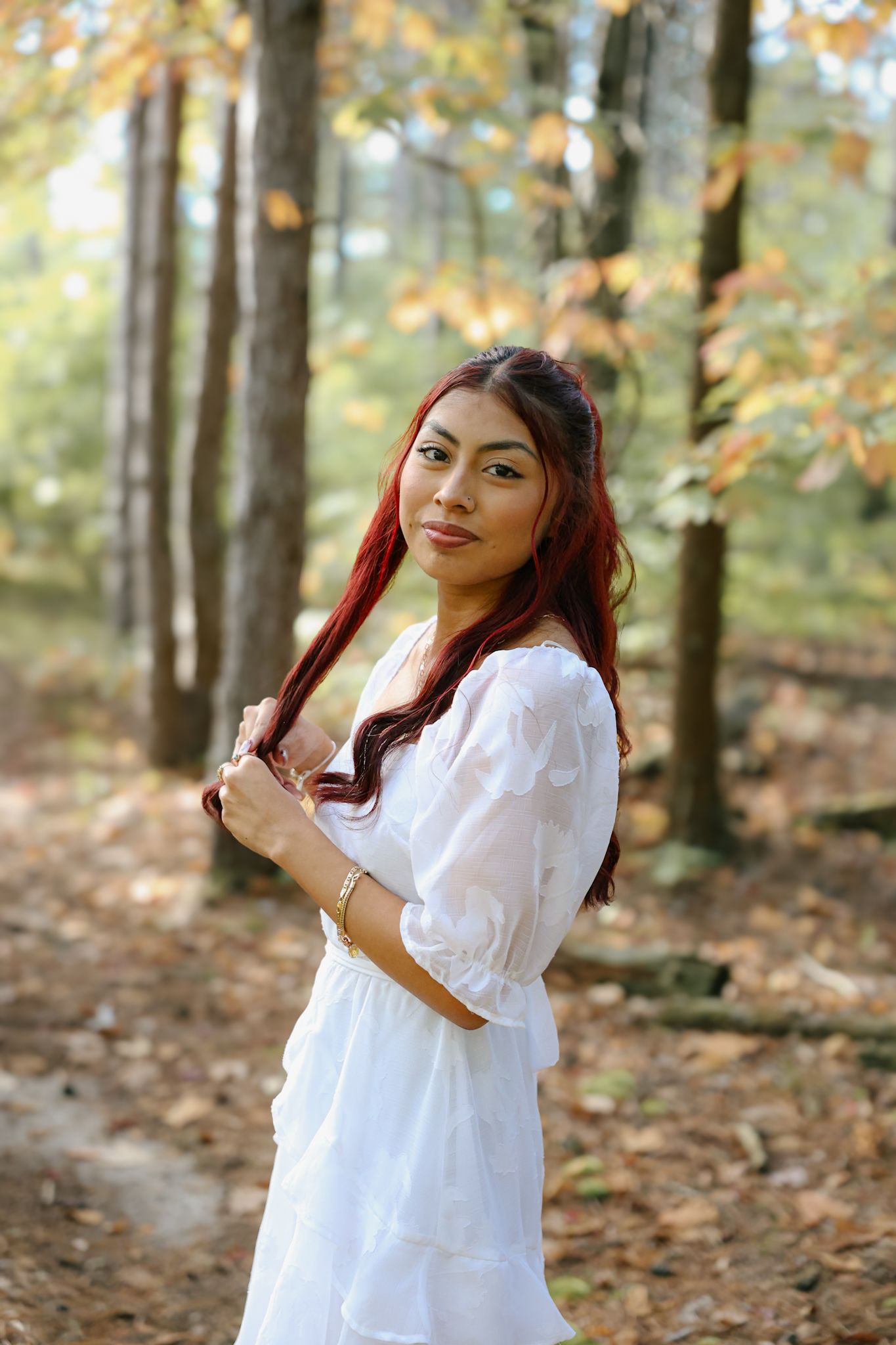 Young woman with red hair in a white dress posing in an autumn forest with colorful leaves.