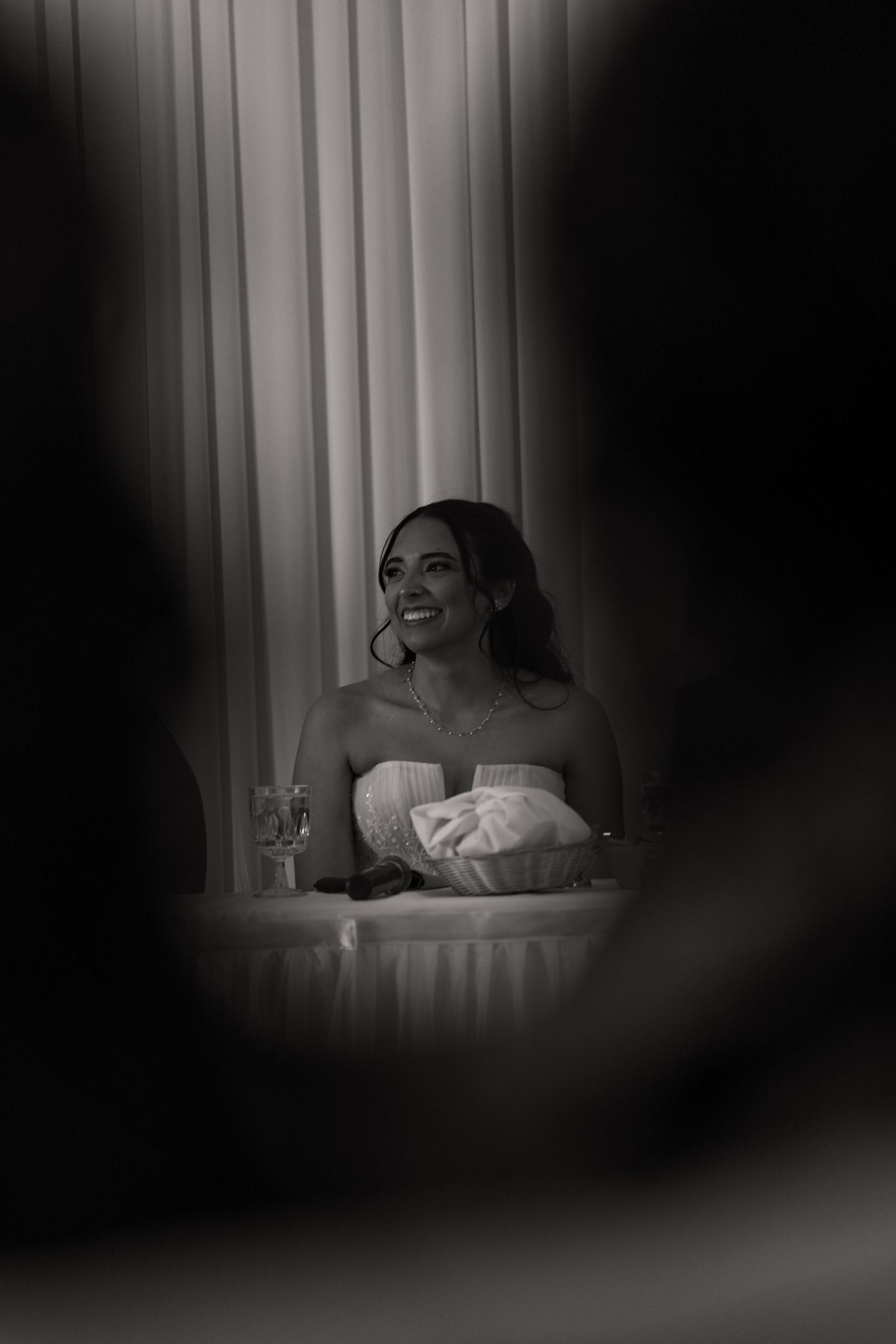 Black and white photo of smiling bride in strapless gown seated at wedding reception table.