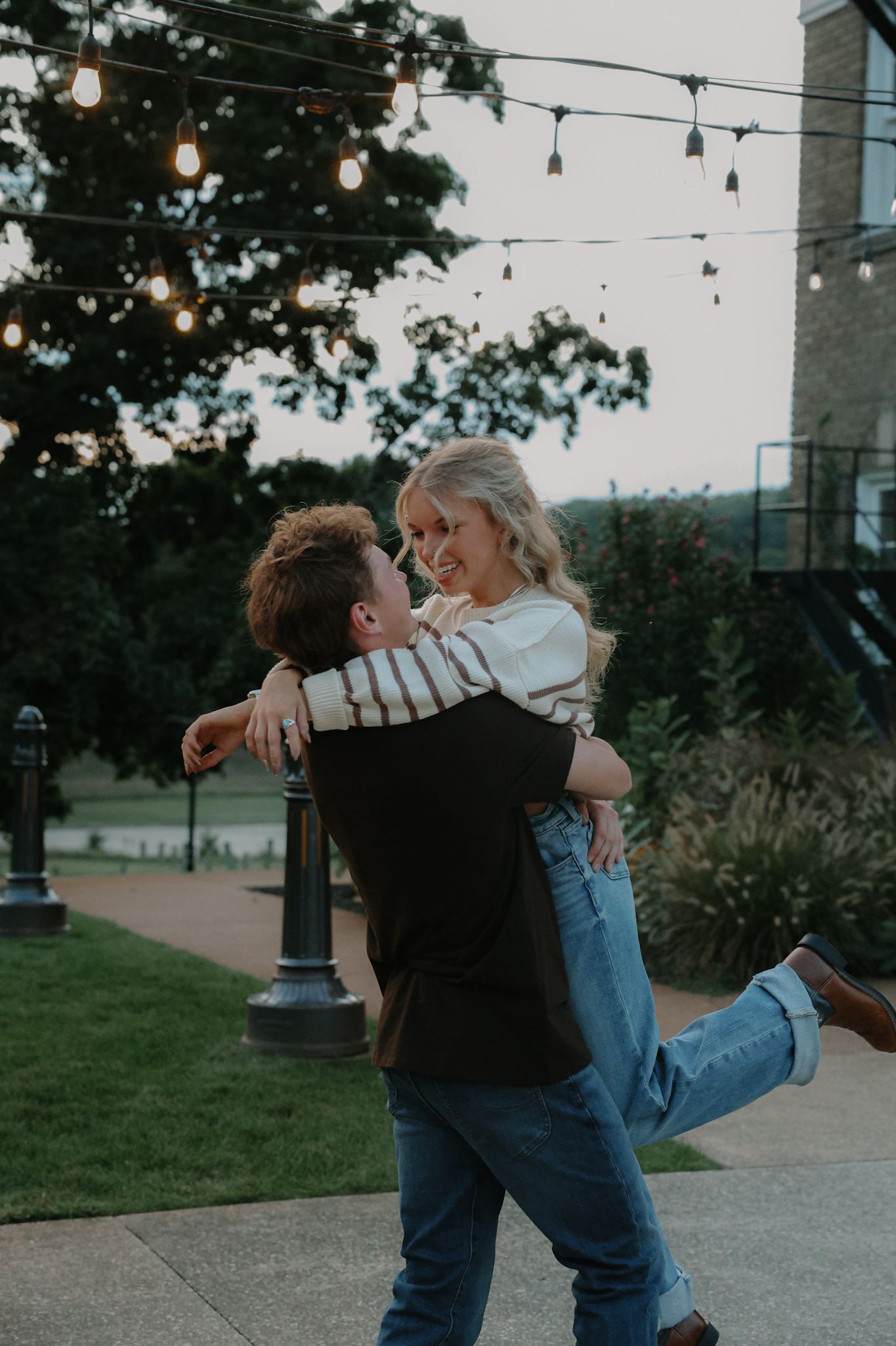 A man lifts a smiling woman in his arms on an outdoor pathway adorned with string lights at dusk.