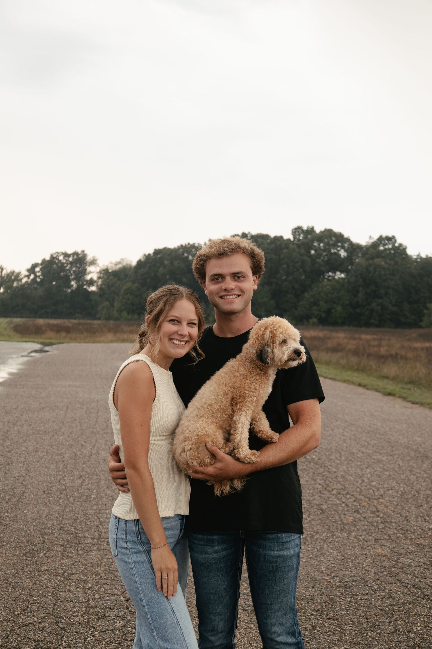A smiling couple stands on a rural road holding a fluffy golden doodle dog with trees in the background.
