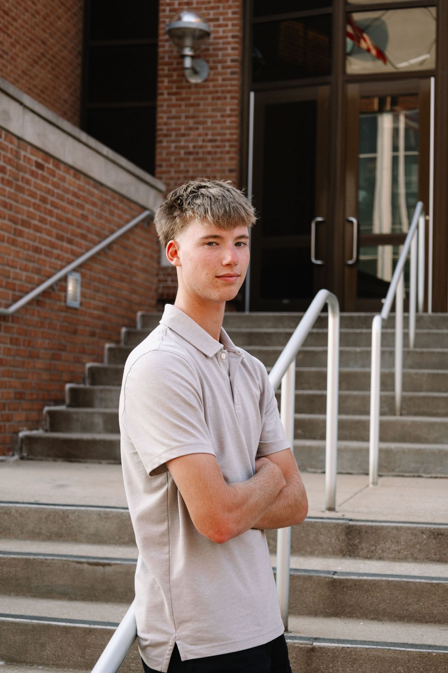 Young man with crossed arms stands confidently on brick building steps wearing a light polo shirt.