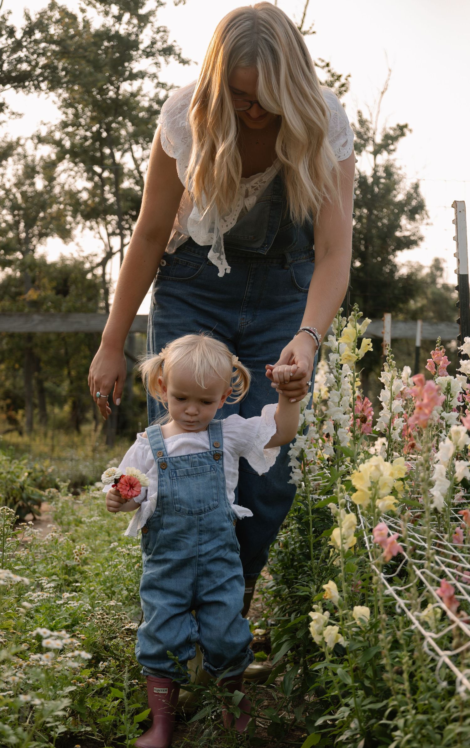 A mother and toddler in matching denim overalls walk through a flower garden together.