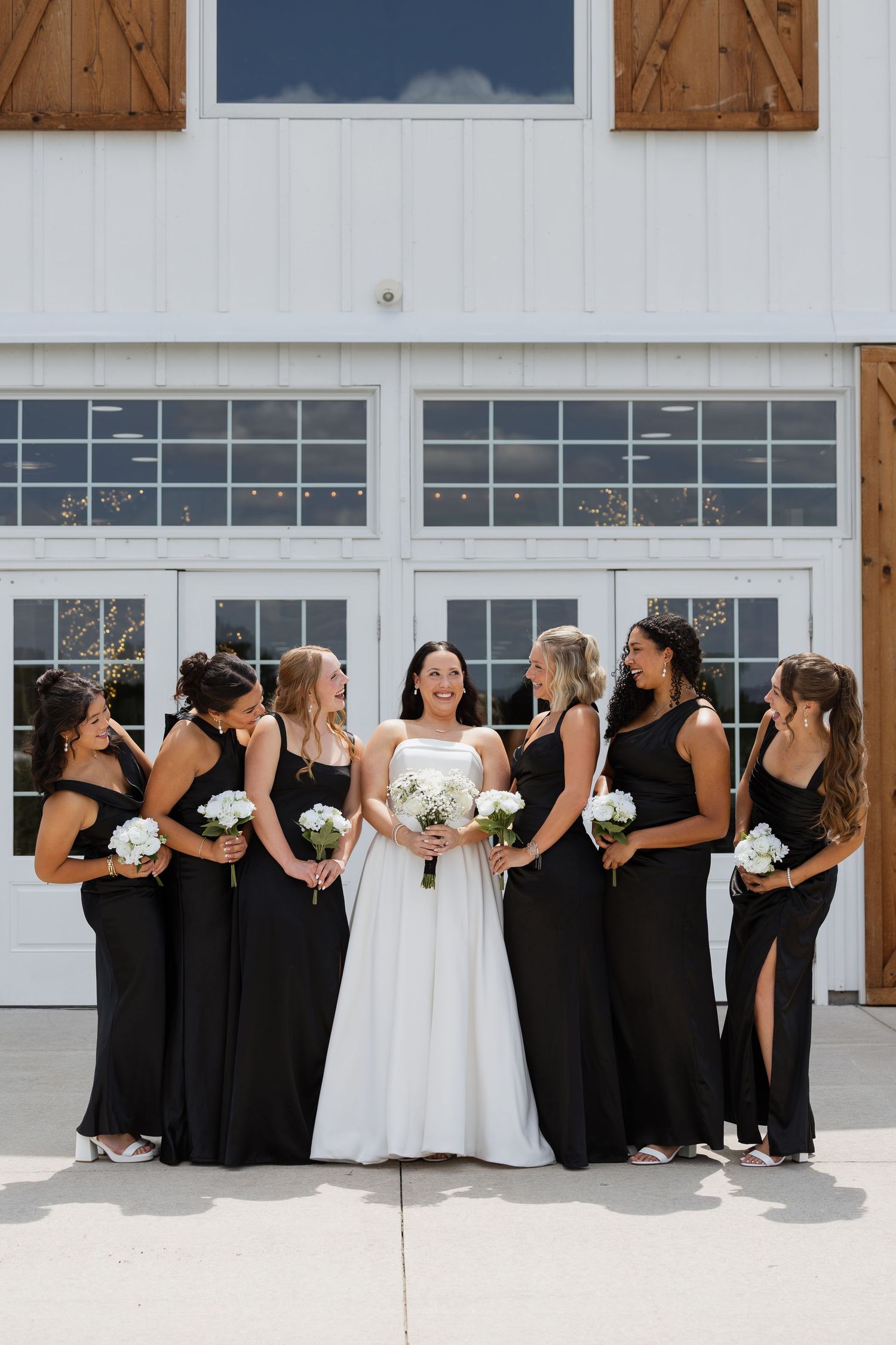 Bride in white gown holds bouquet surrounded by bridesmaids in black dresses outside a white barn venue.