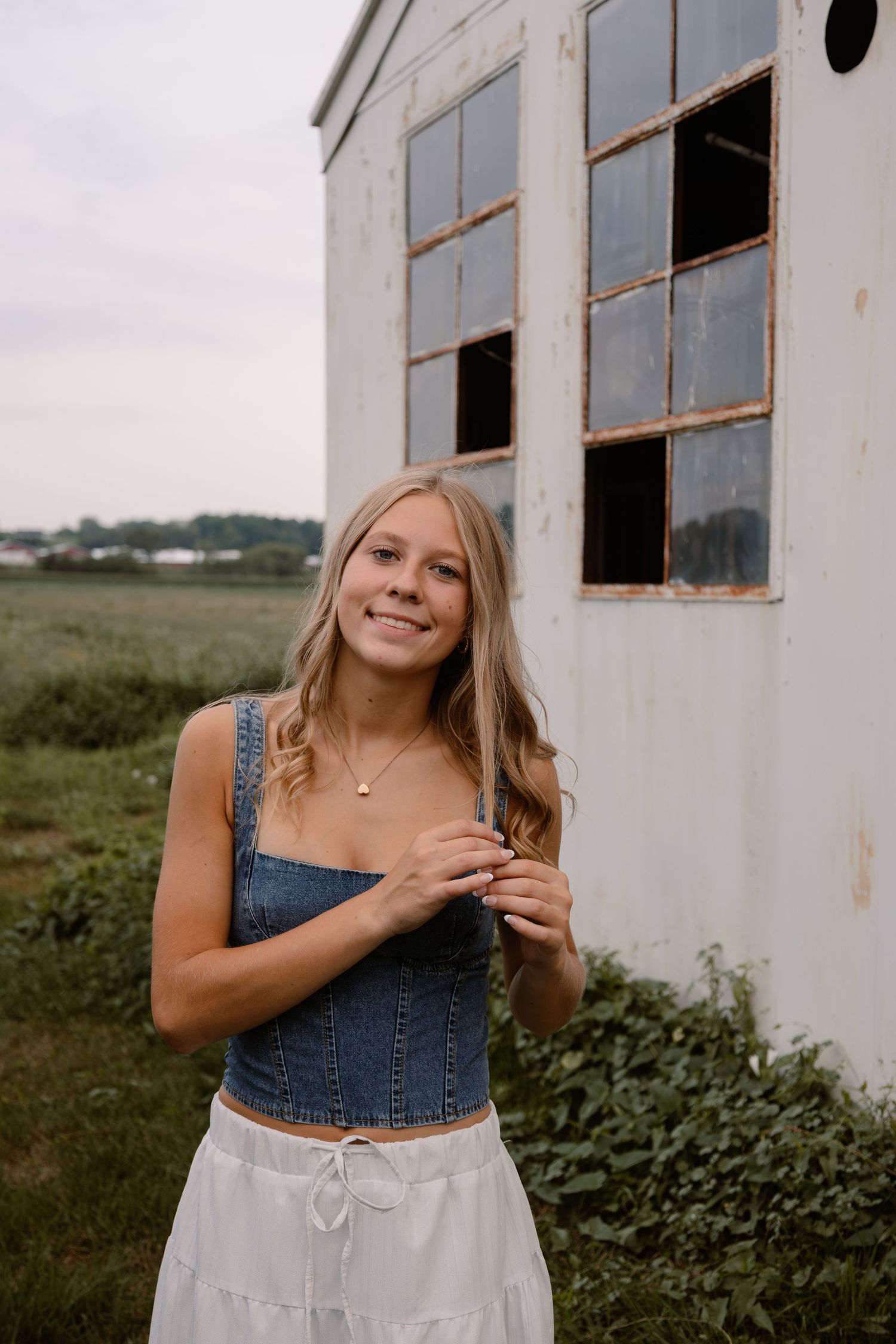 Young woman in denim crop top and white skirt smiling outdoors near a rustic white barn.