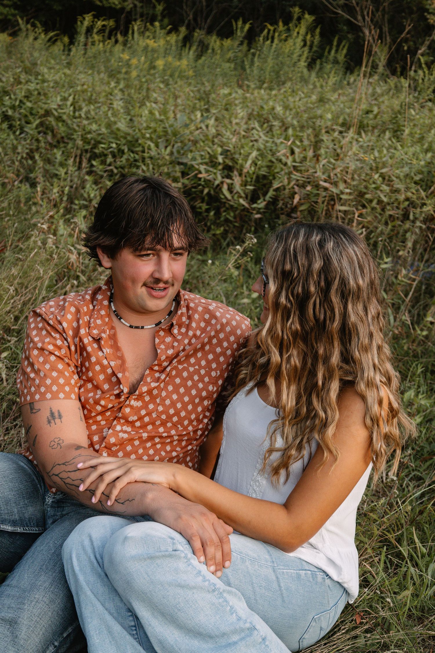 A couple sits together in a grassy field, the man in a polka dot shirt smiling at a woman with wavy hair.