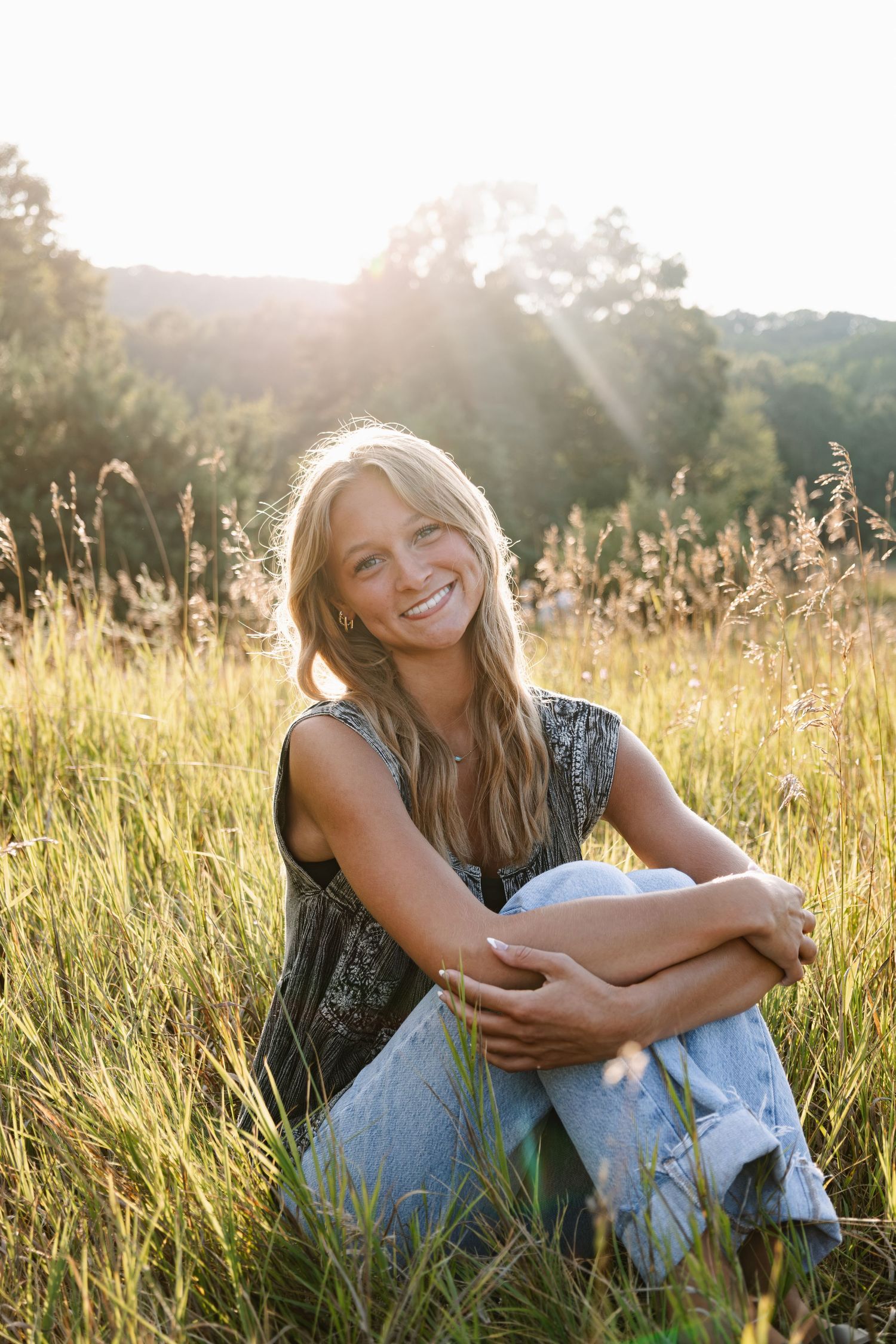 Smiling young woman sitting in a sunlit grassy field, wearing jeans and a sleeveless top, with trees in the background.