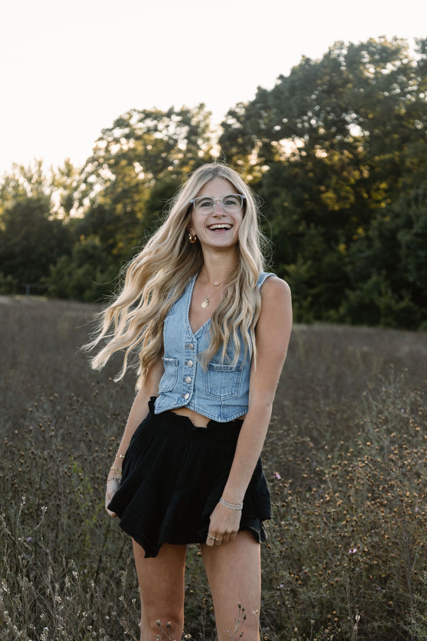 Smiling blonde woman in denim vest and black skirt standing in a sunlit field with trees in the background.