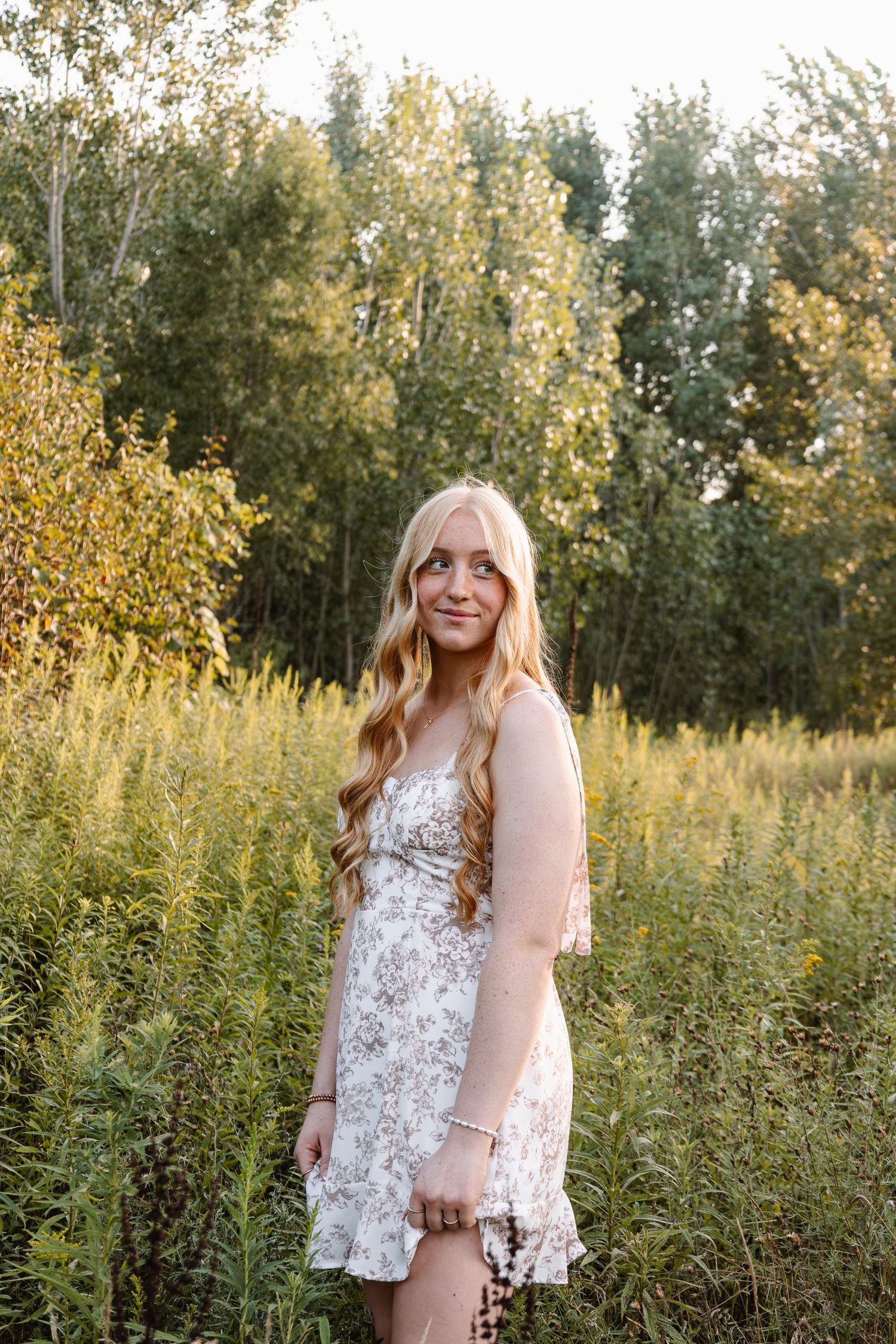 Young blonde woman in floral sundress posing in a lush green meadow with trees in golden hour light.