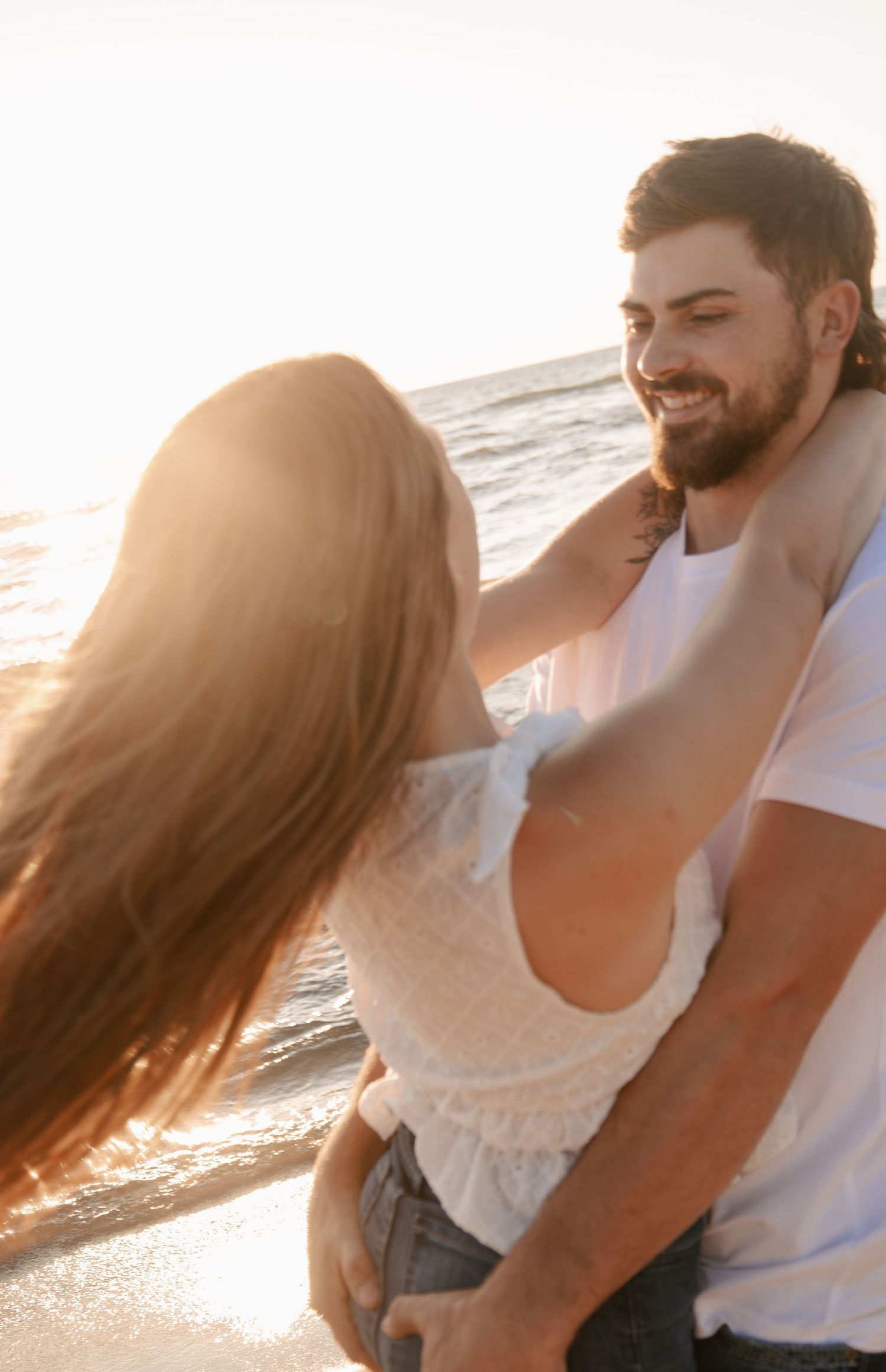 Smiling bearded man embracing woman on sunny beach at golden hour sunset.