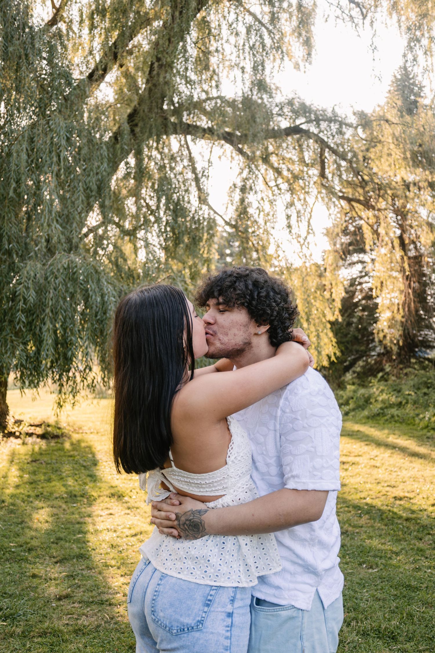 A couple shares a romantic kiss outdoors beneath a weeping willow tree on a sunny day.