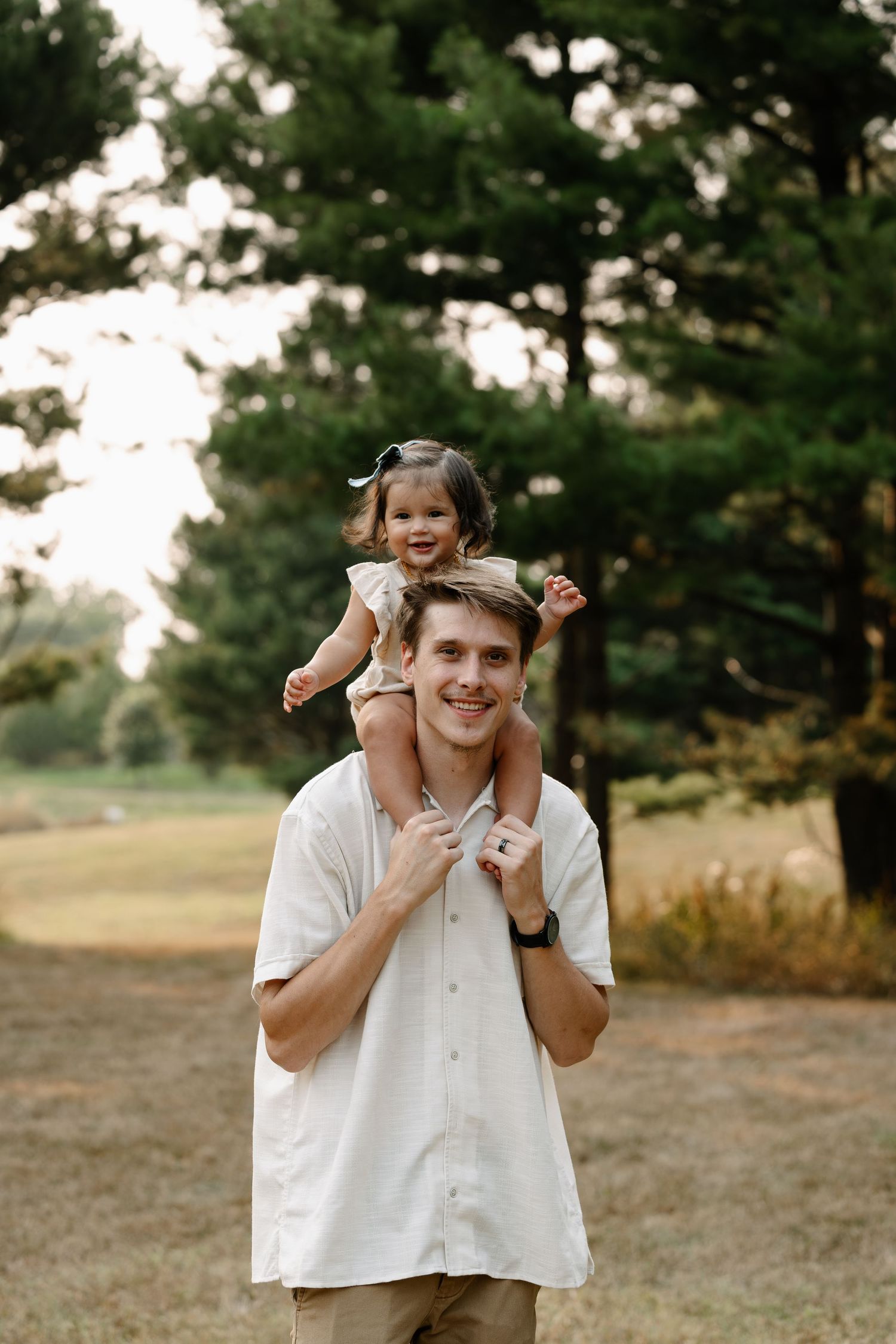 A smiling man carries a happy toddler girl on his shoulders outdoors in a lush green park setting.