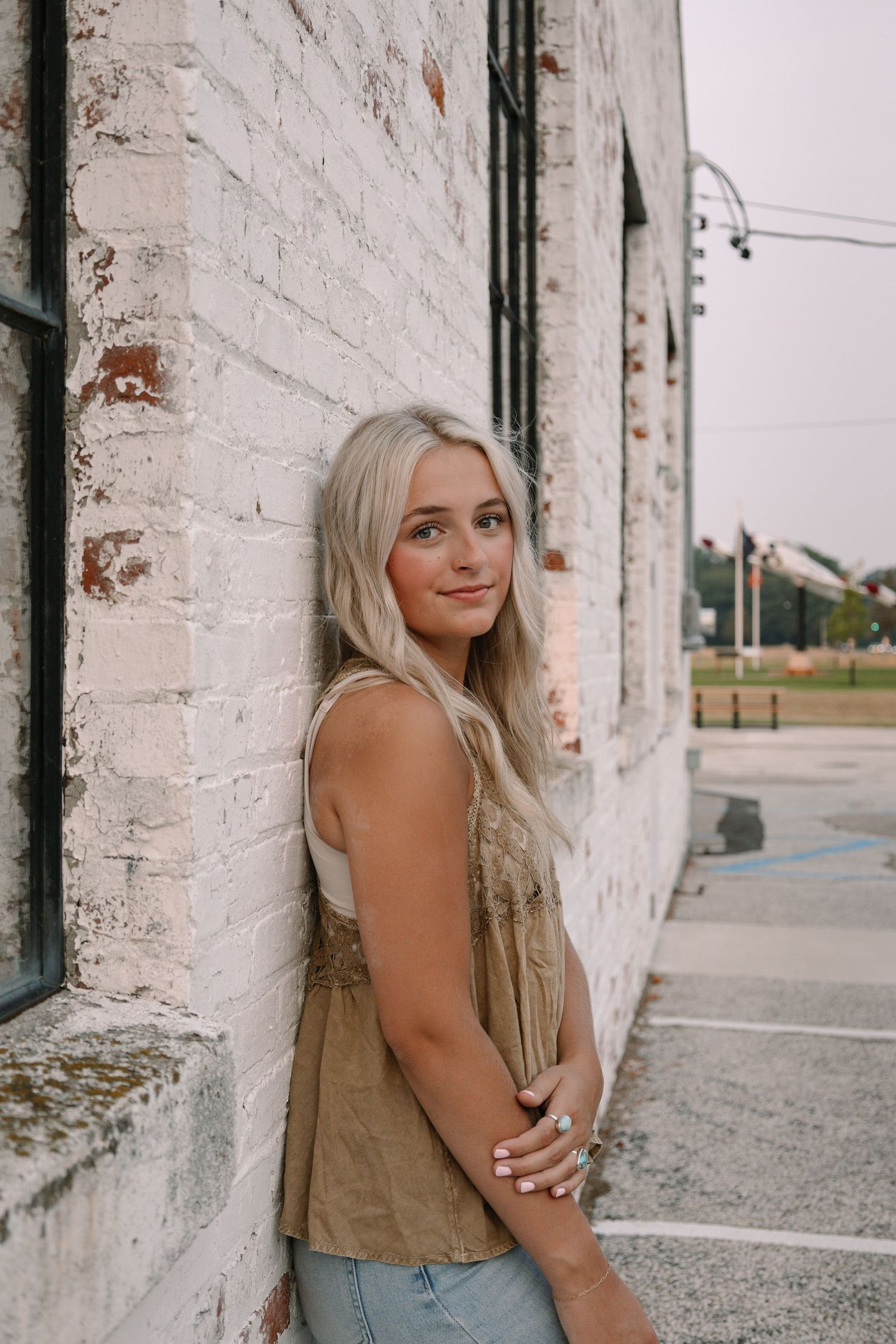Young blonde woman in olive tank top leaning against white brick wall, smiling over shoulder outdoors.