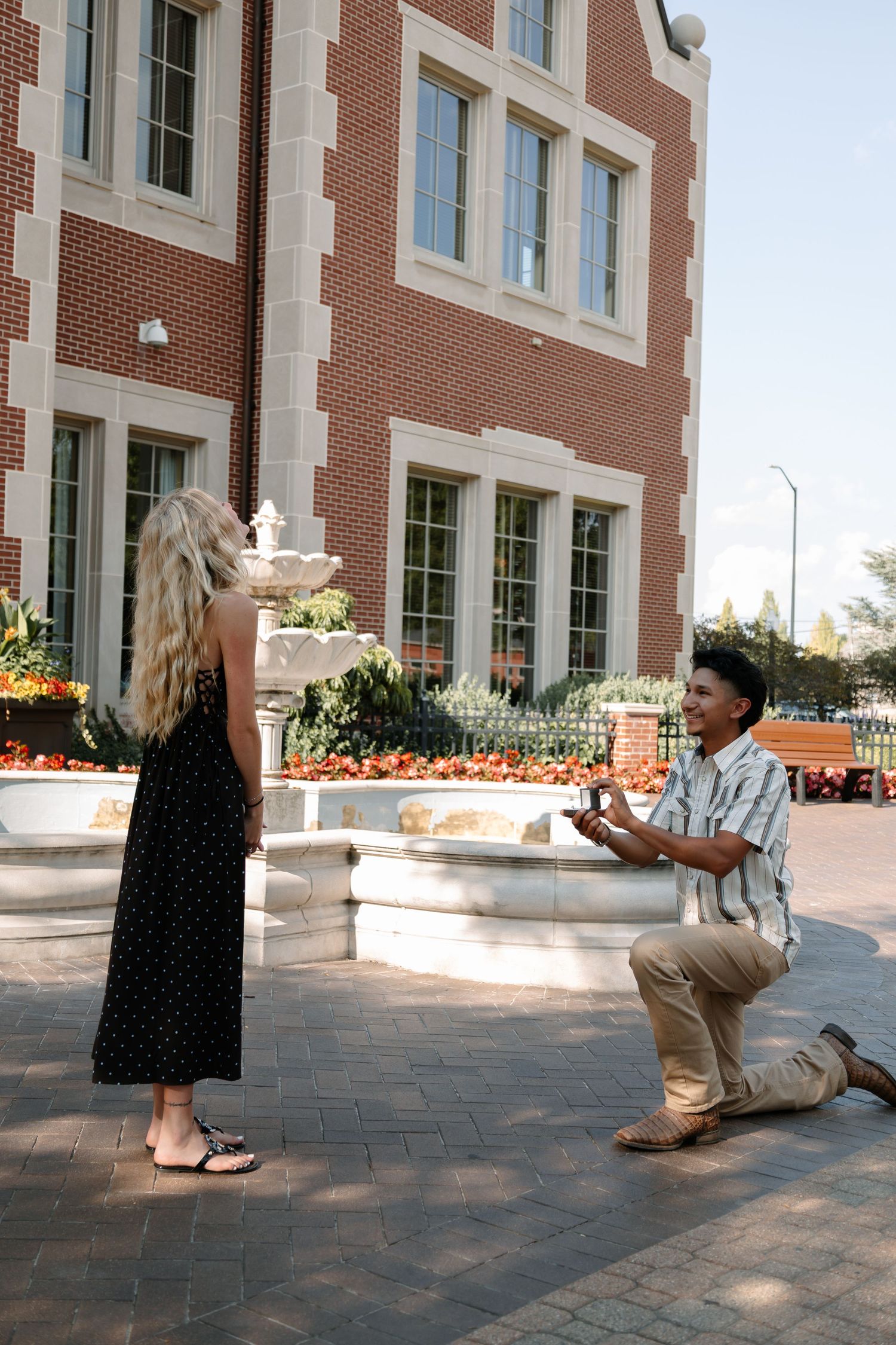 Man proposes on one knee to woman outside brick building with fountain.