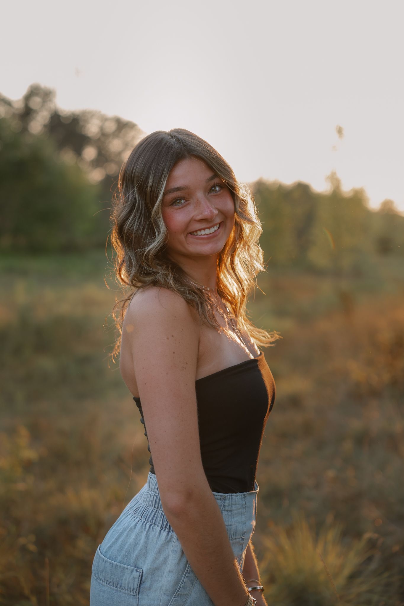 Young woman in black tube top and light jeans smiling in a golden hour field.