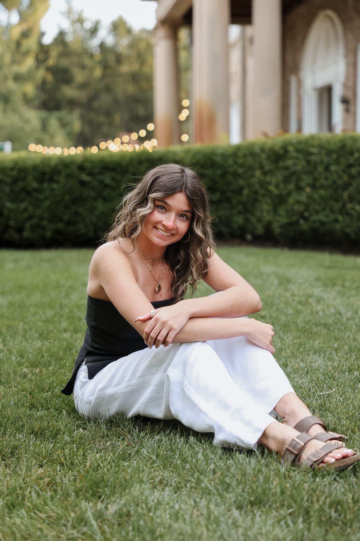 Smiling young woman sitting on green grass in black top and white pants at an outdoor garden venue.