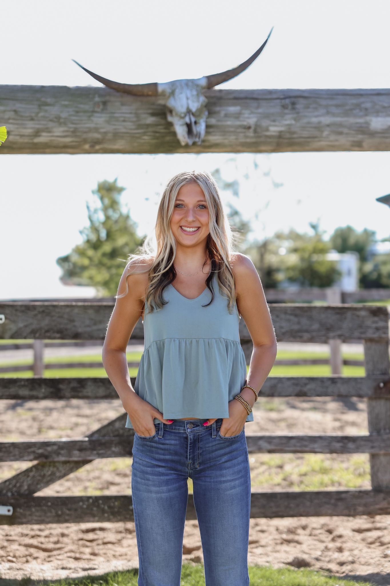 Smiling blonde woman in teal tank top and jeans standing by a rustic fence with a steer skull.