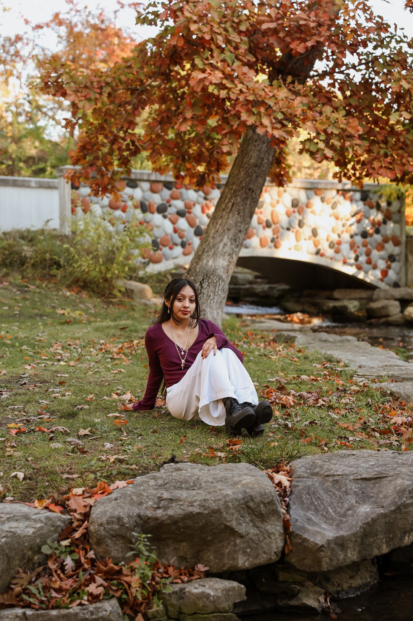 Young woman in burgundy top and white skirt sitting near rocks in colorful autumn park.