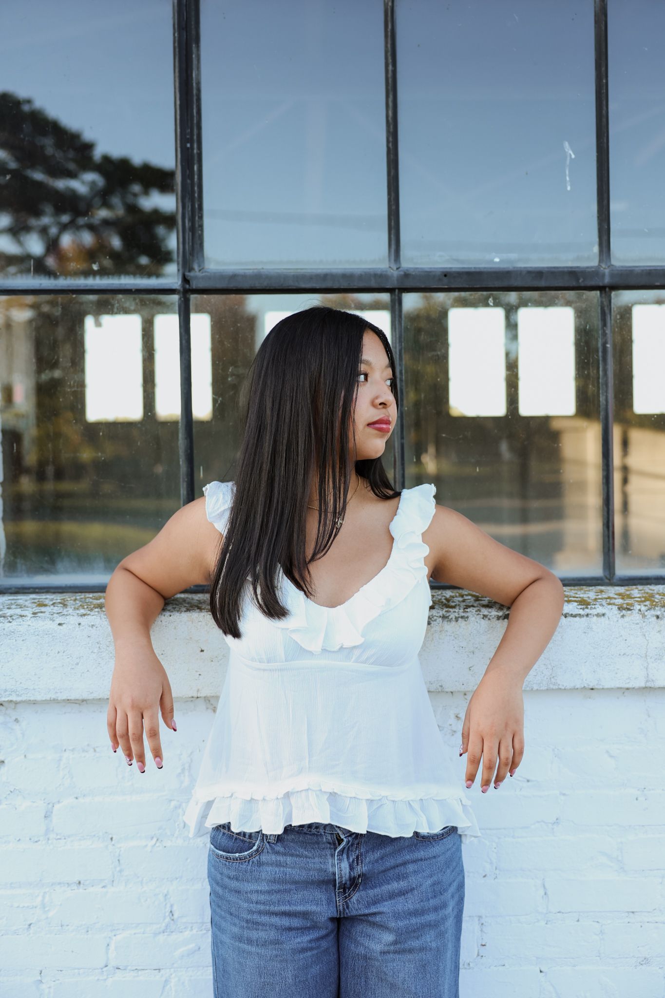 Young Asian woman in white ruffle tank top leans against a white brick wall near large windows.