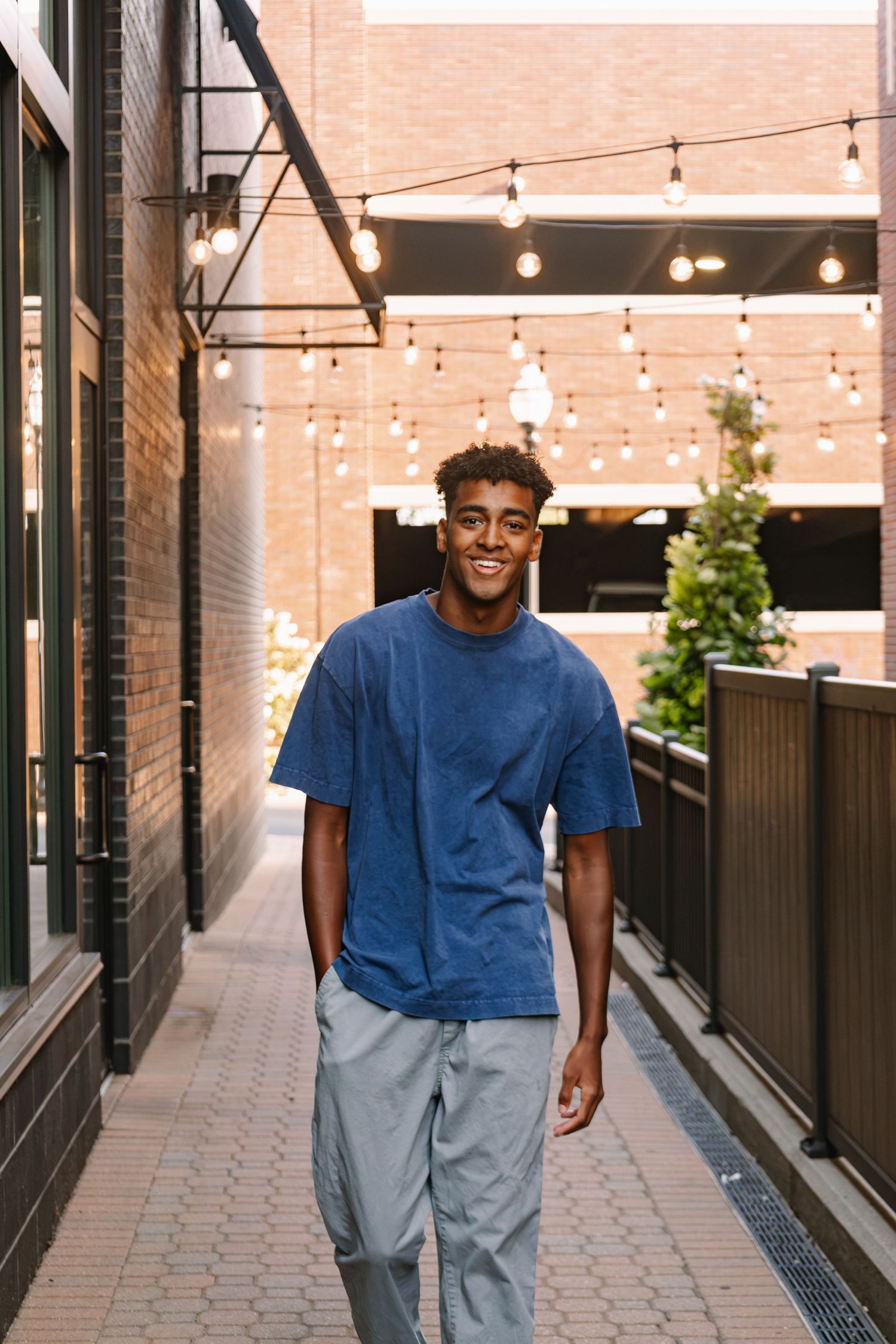 Smiling young Black man in blue oversized t-shirt and gray pants walking through urban brick alleyway with string lights.
