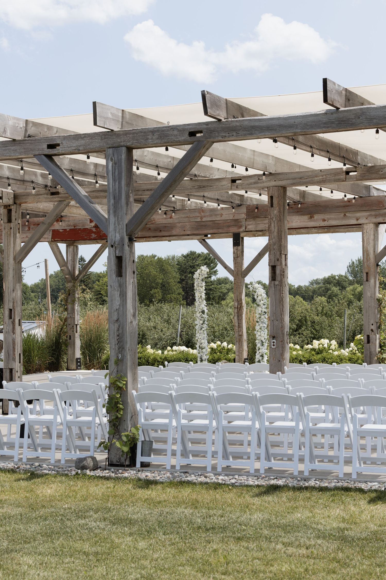 Outdoor wedding ceremony setup with white folding chairs under a rustic wooden pergola surrounded by lush greenery.