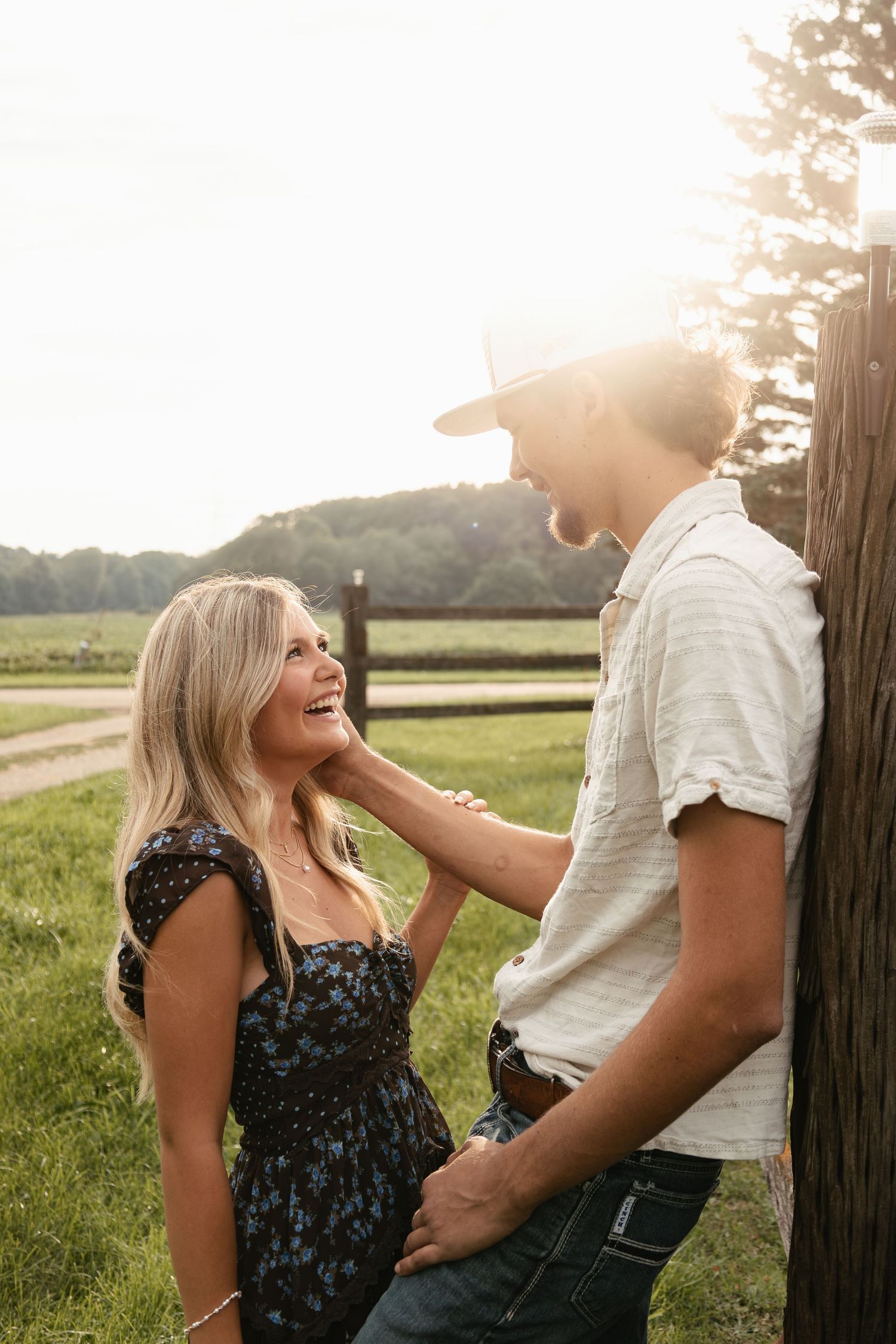 A couple shares a romantic moment at golden hour by a wooden fence on a green farm, the woman smiling up at the man in a white hat.