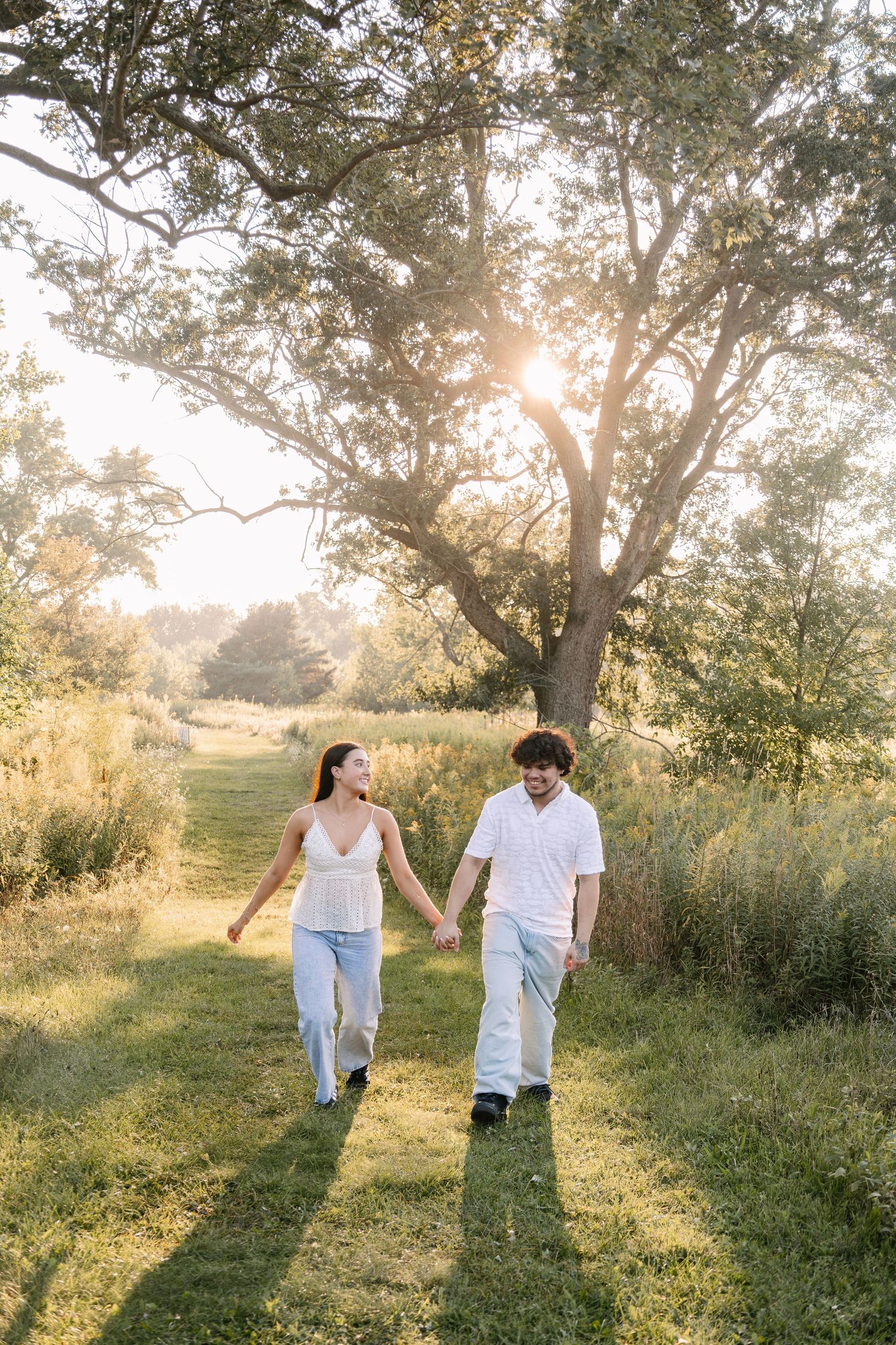 A couple holds hands walking through a sunlit green meadow path with large trees glowing in golden hour light.