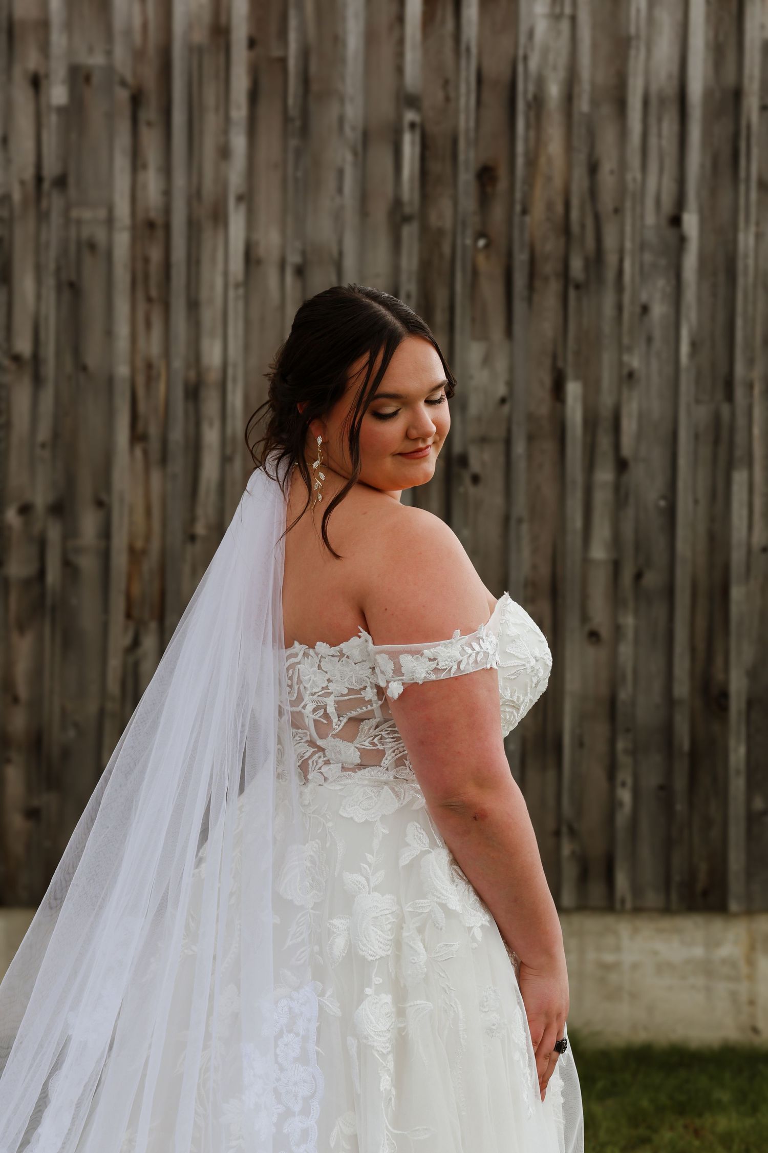 Beautiful bride in off-shoulder lace wedding gown with veil posing in front of rustic wooden barn wall.