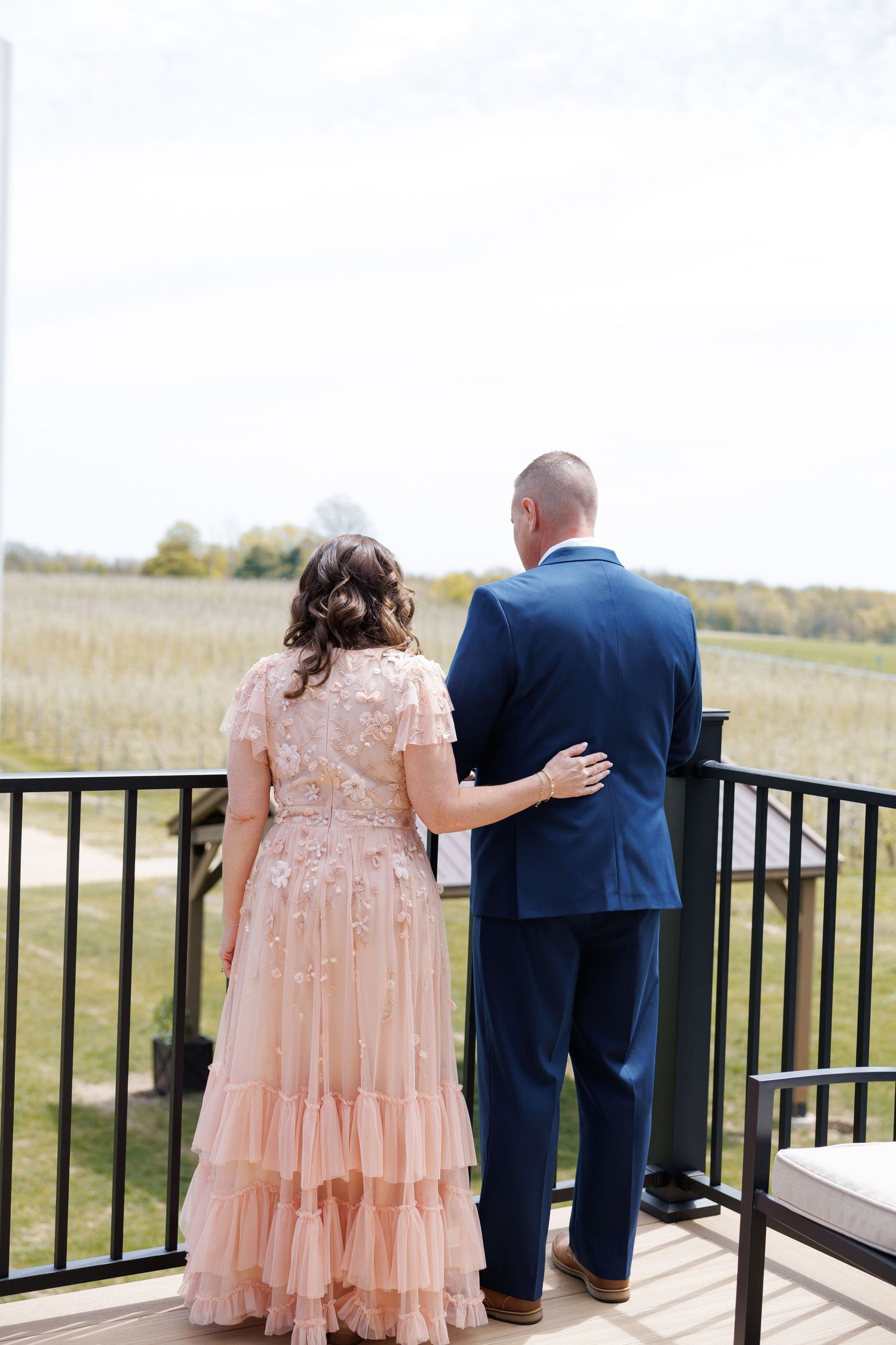 Bride in pink floral gown and groom in navy suit stand together overlooking a scenic cornfield.