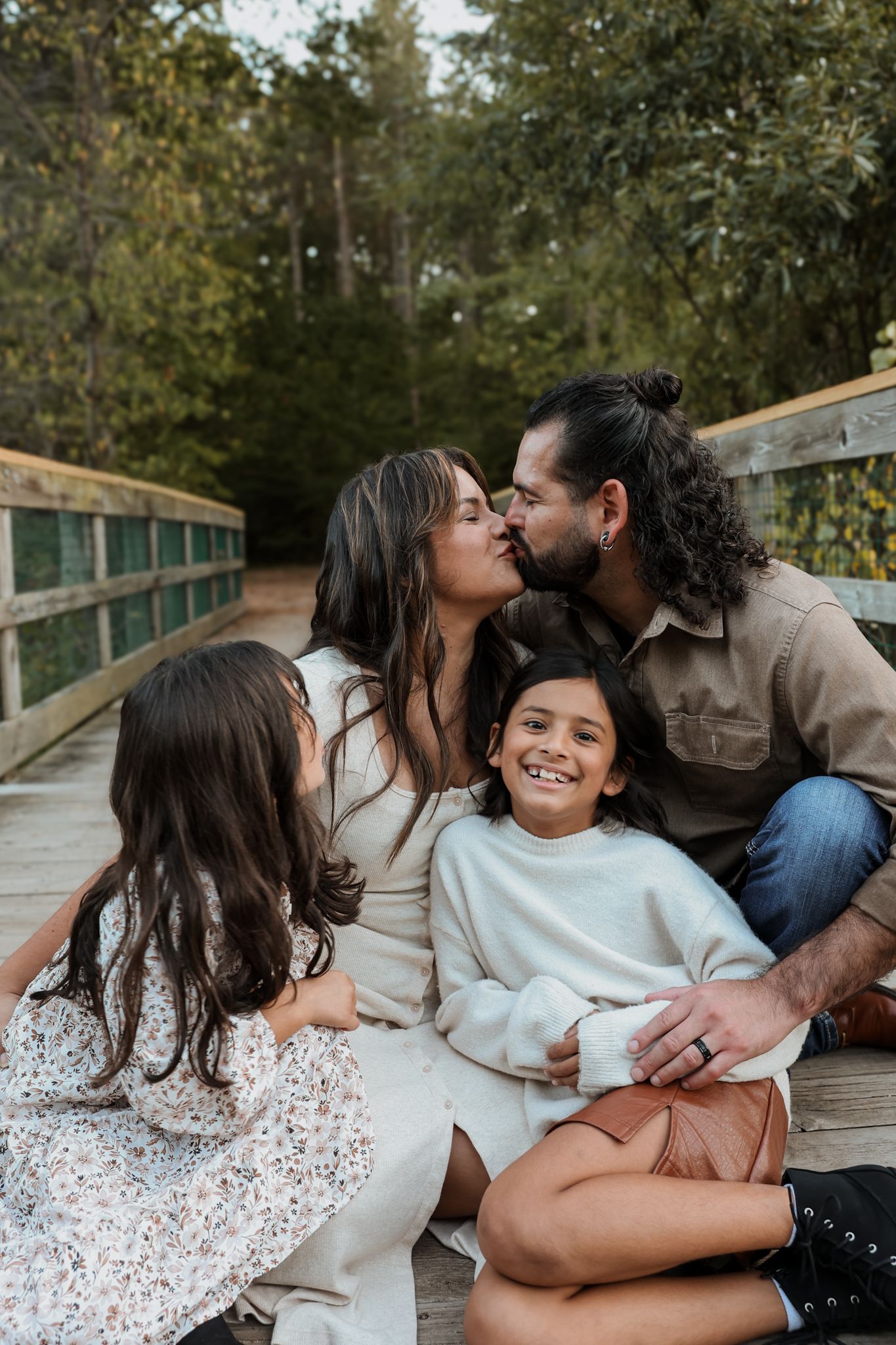Family sits on wooden bridge steps as parents kiss while daughters smile and cuddle.